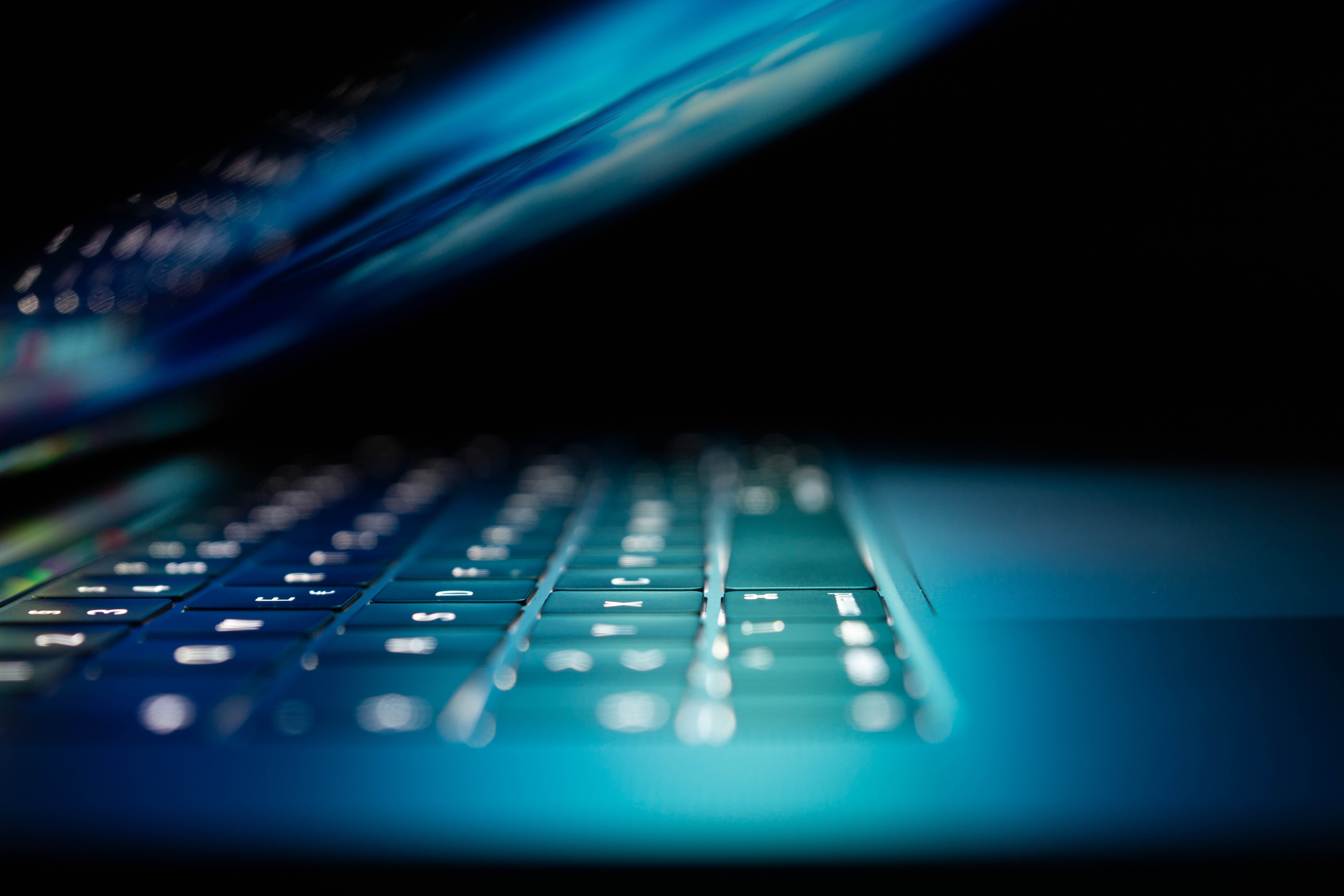 A laptop computer, seen from the side, is slightly open and glows with blue light in a dark room.
