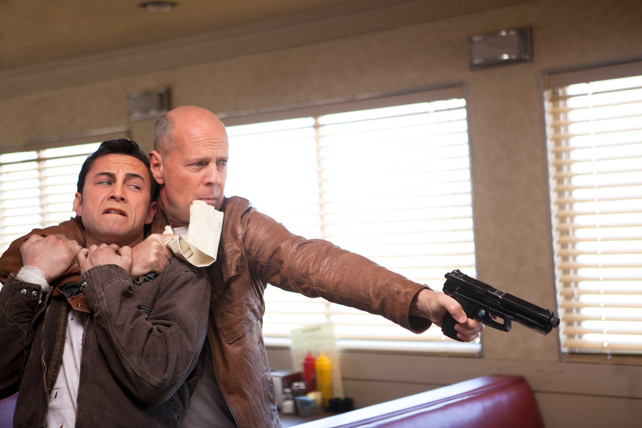A man in a diner holds another man hostage in a chokehold while he points a gun at someone off camera. 