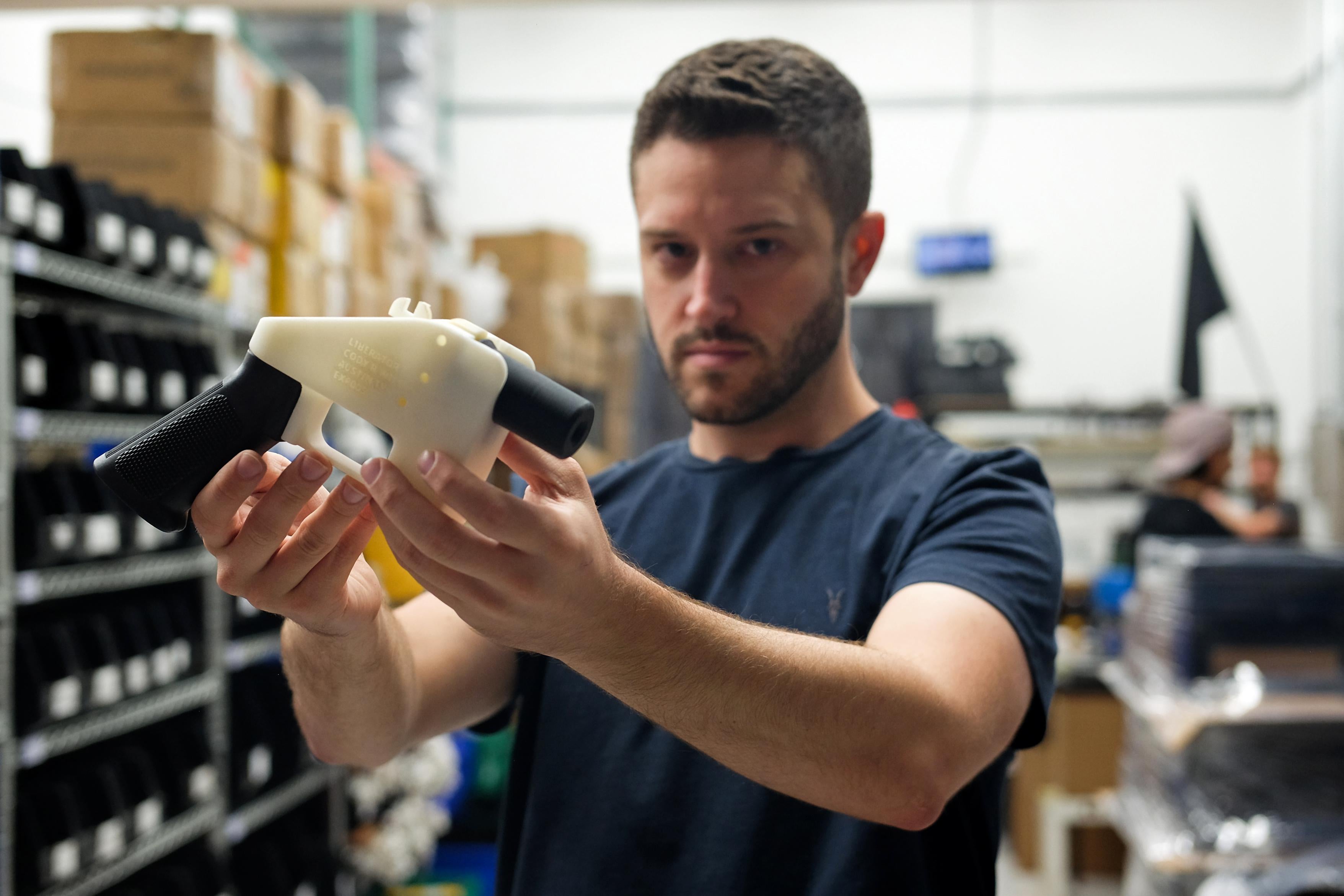 Cody Wilson holds up a black-and-white 3D-printed plastic gun while standing in his factory.