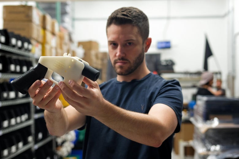 Cody Wilson holds up a black-and-white 3D-printed plastic gun while standing in his factory.