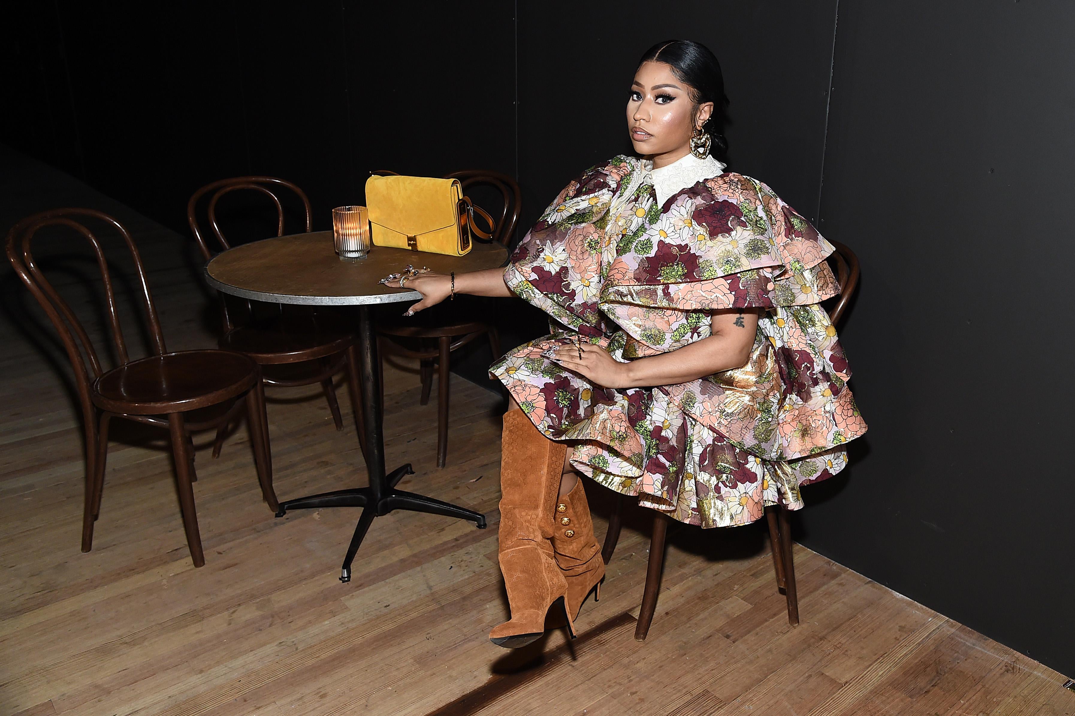 Nicki Minaj, in an elaborate dress and boots, sits at a table that holds a purse and a candle.