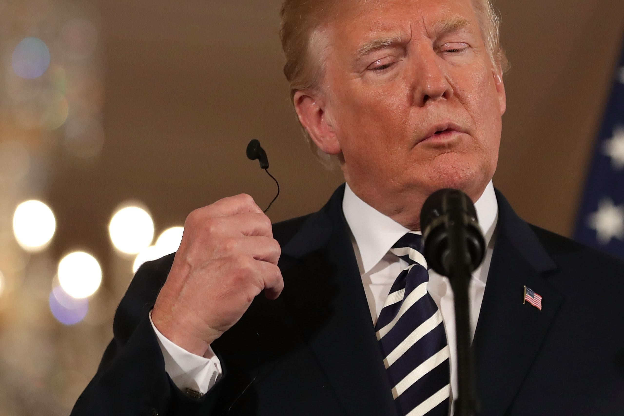 President Trump, standing at a podium, pulls an audio ear bud out of his ear during a news conference. 
