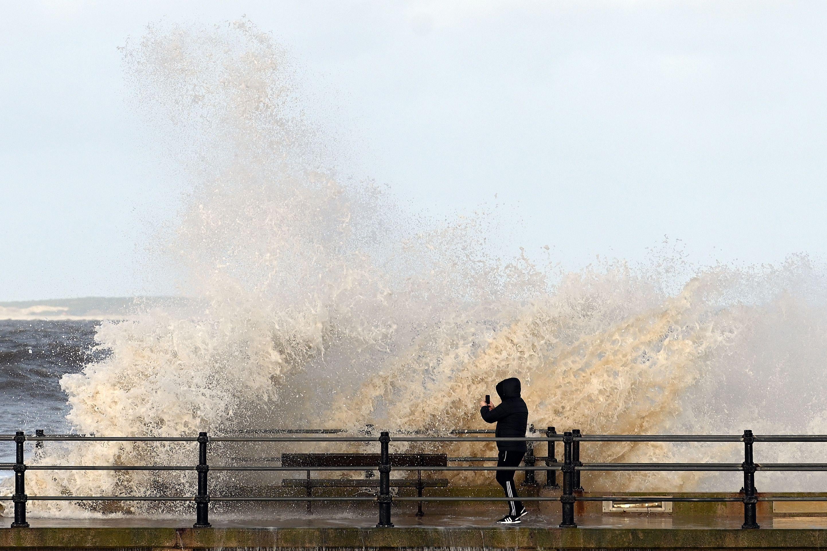 A man stands on a sea wall as waves break overhead.
