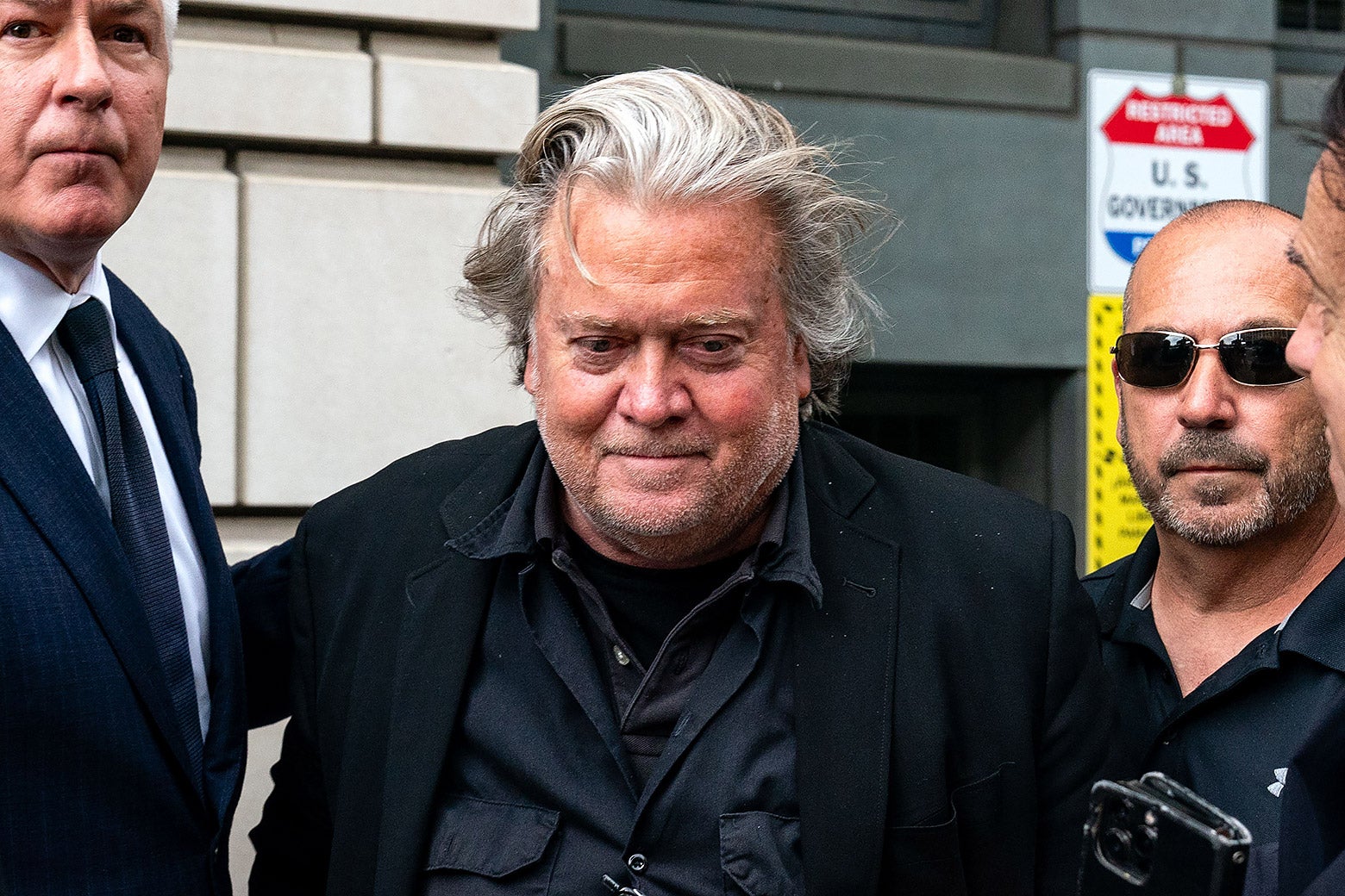 Bannon staring into space, looking slightly downward, as he's escorted by goons.