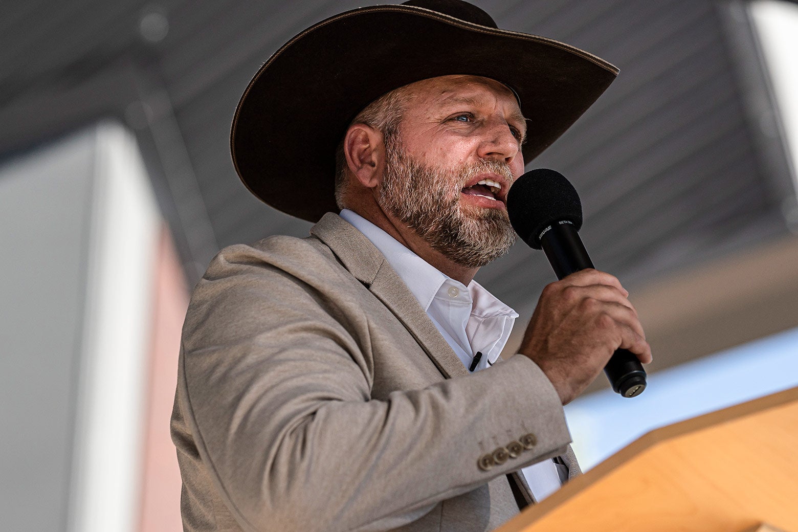 A bearded Bundy, wearing a light-colored suit and a cowboy hat, speaks into a handheld microphone.