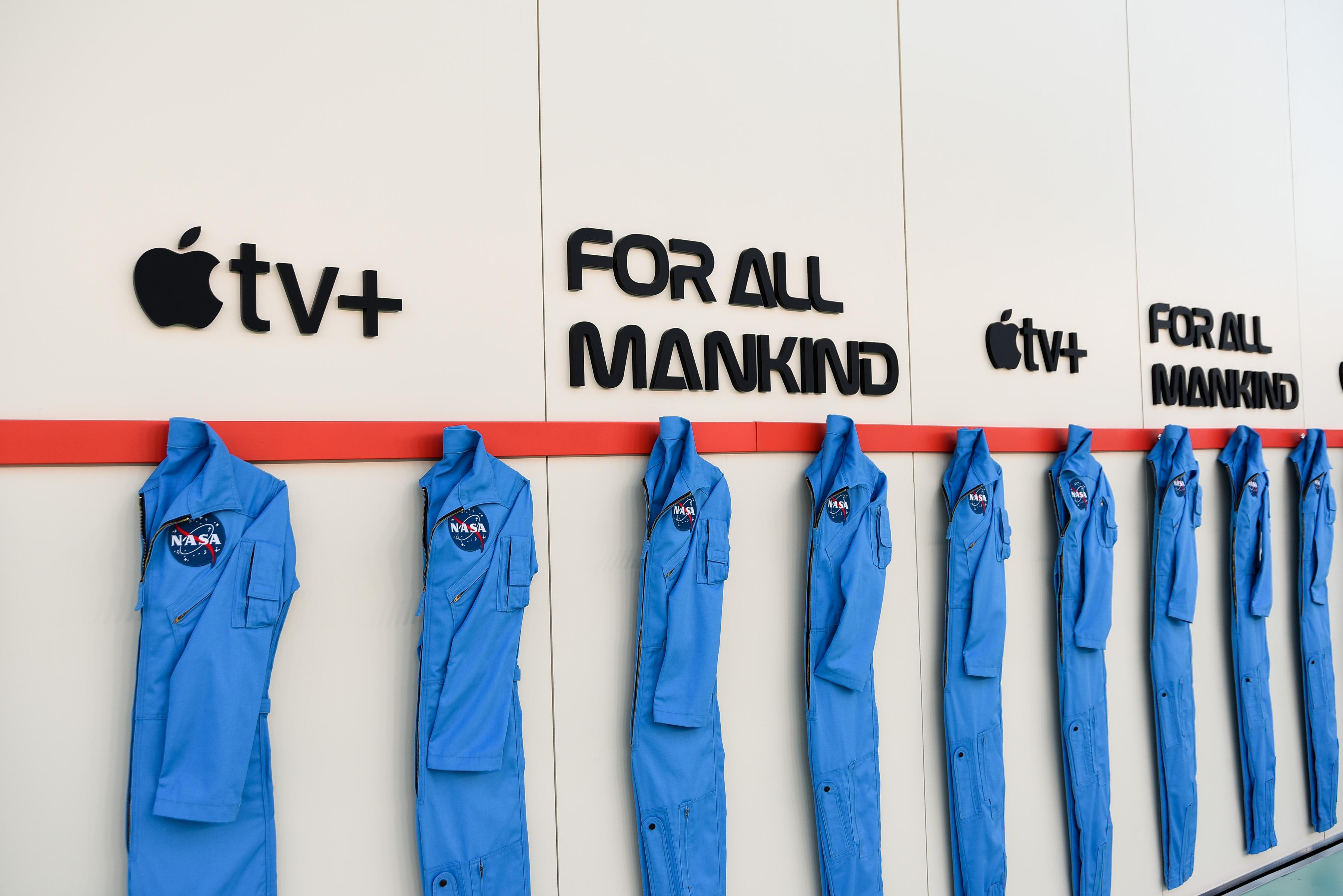 Blue NASA uniforms hung in a line on the wall, under branding for Apple TV+ and For All Mankind. 