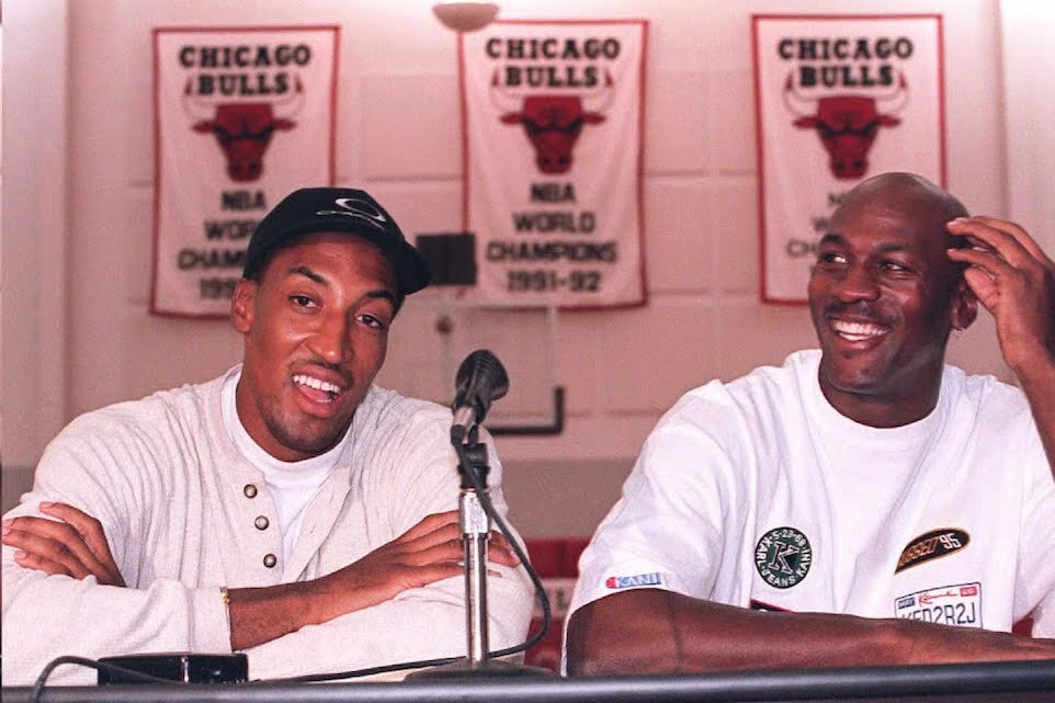 Scottie Pippen and Michael Jordan sitting at a table with a microphone, and behind them are banners for three Bulls championships.