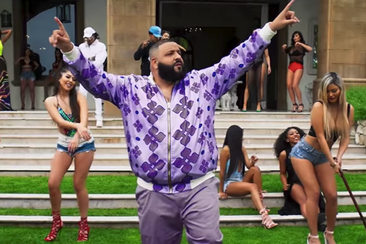 DJ Khaled points both index fingers in the air as scantily clad women dance behind him.