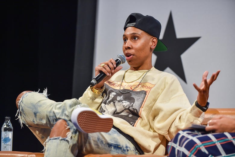 A woman in an oversized yellow sweatshirt, a backward navy blue baseball cap, and ripped blue jeans sits on a couch onstage. She is holding a microphone and addressing the audience.