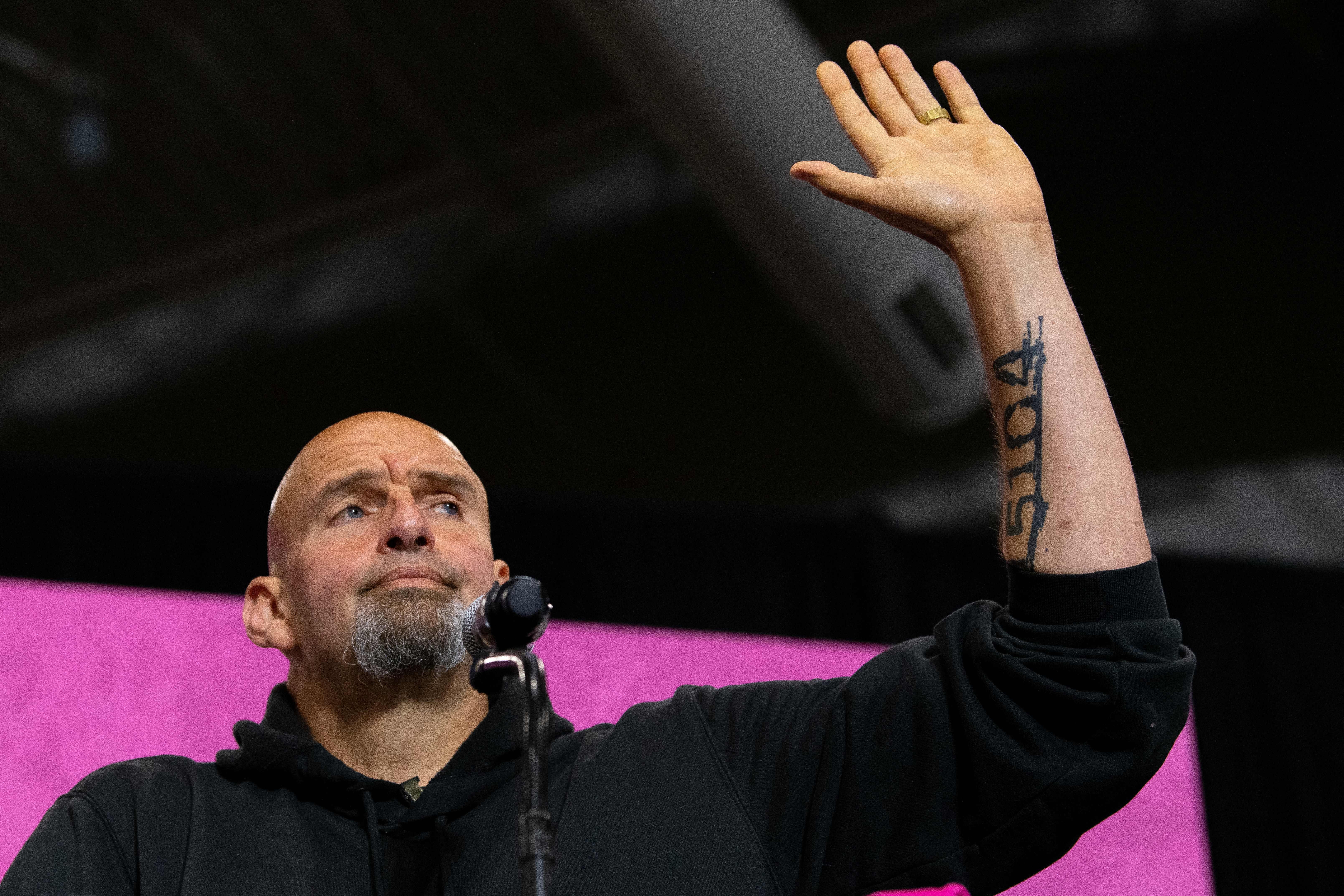 John Fetterman stands in front of a mic and waves.