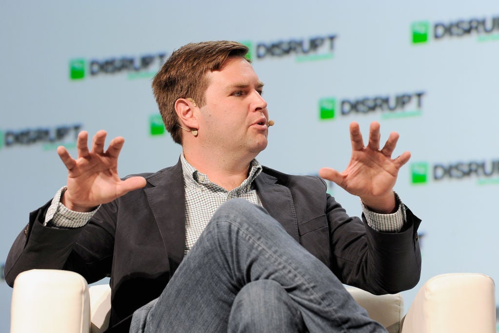 J.D. Vance, wearing a sport coat and a wireless headset, gestures while seated in a plush chair at a tech conference.