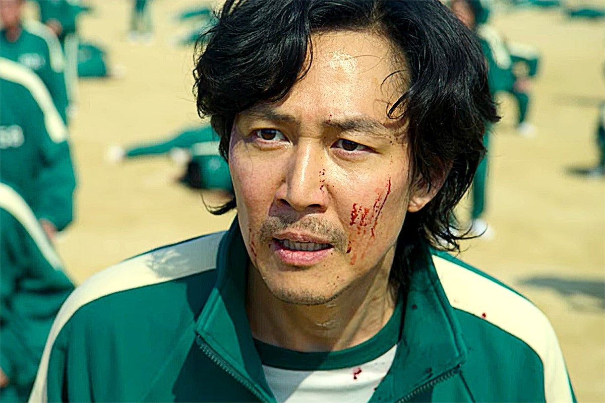 Gi-hun with dried blood on his left cheek stands in a green tracksuit amid other players in green tracksuits