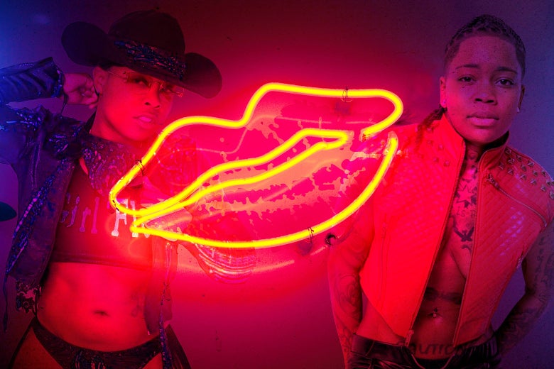 Dancers Girl Flexxx and Kaution in their stripper attire against a neon sign of lips.