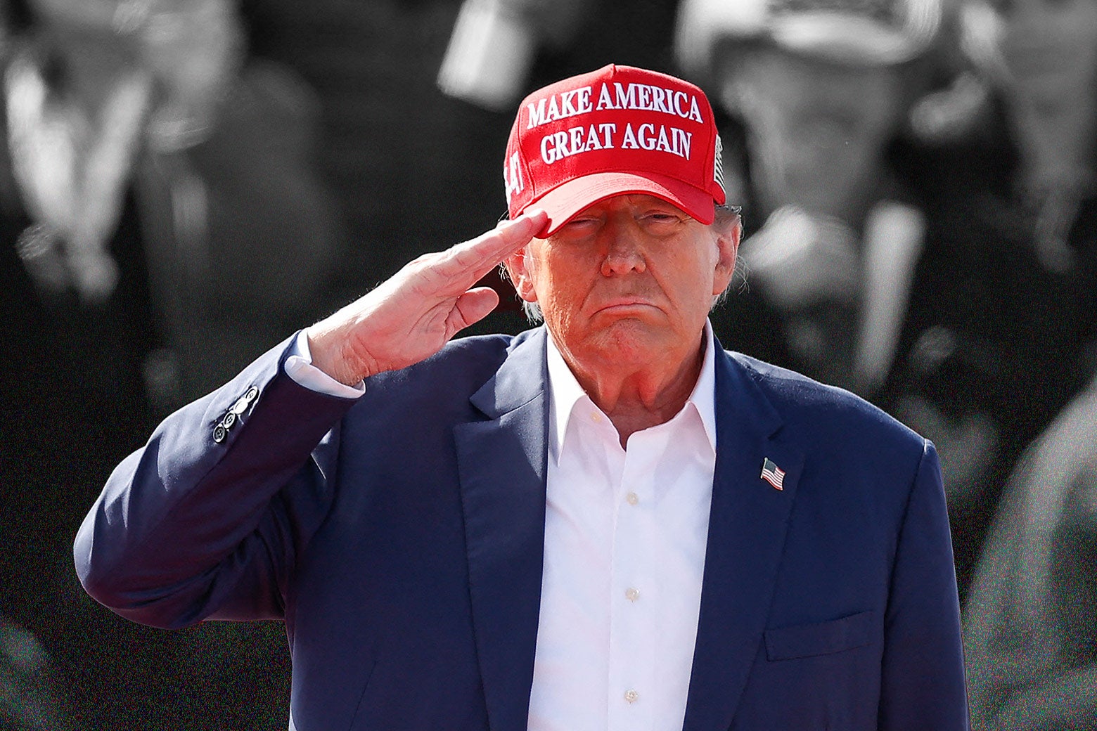 Trump saluting and frowning, in a blue suit with no tie, and a red MAGA hat.