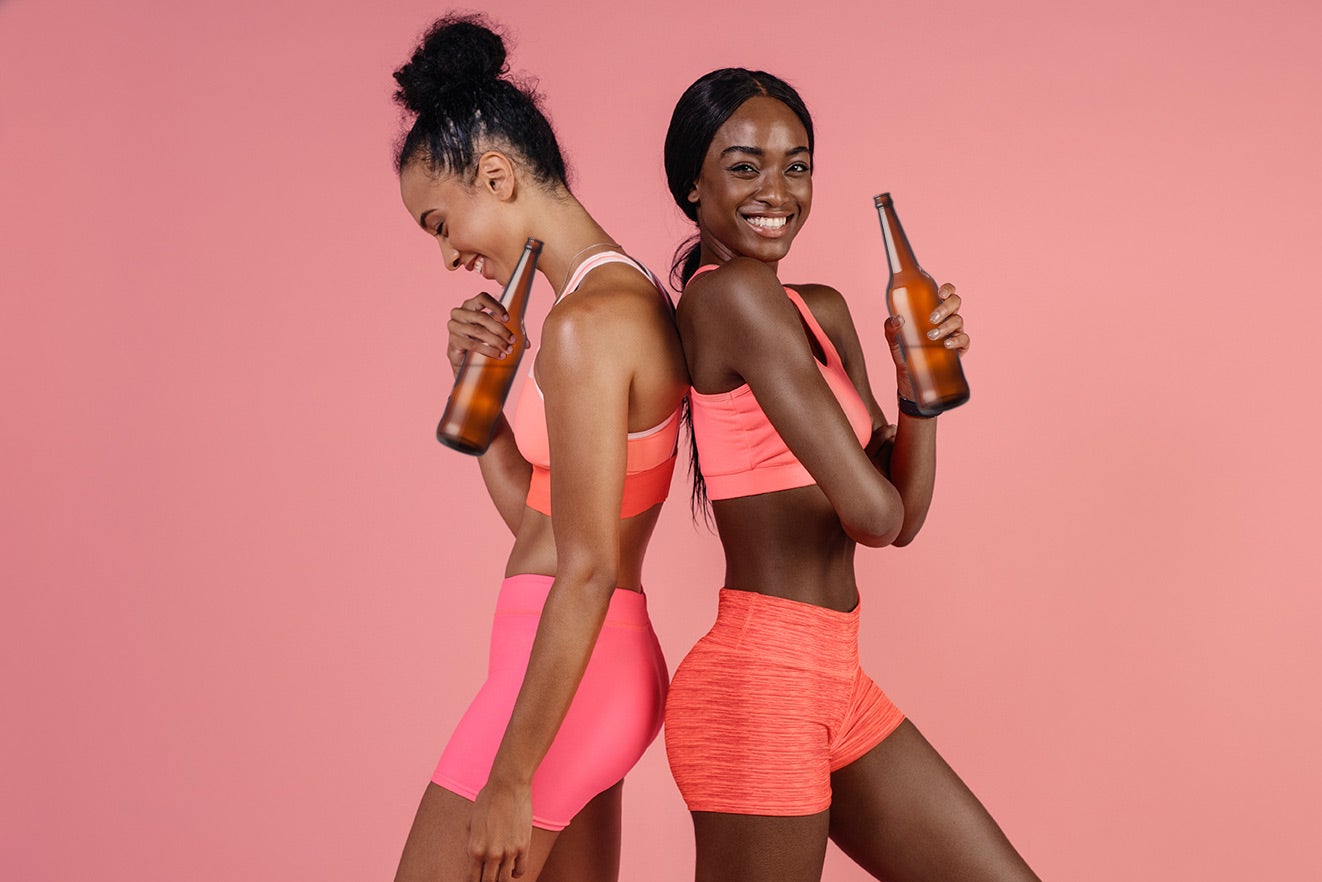 Two girls in fitness outfits hold beer bottles. They love to work out drunk.