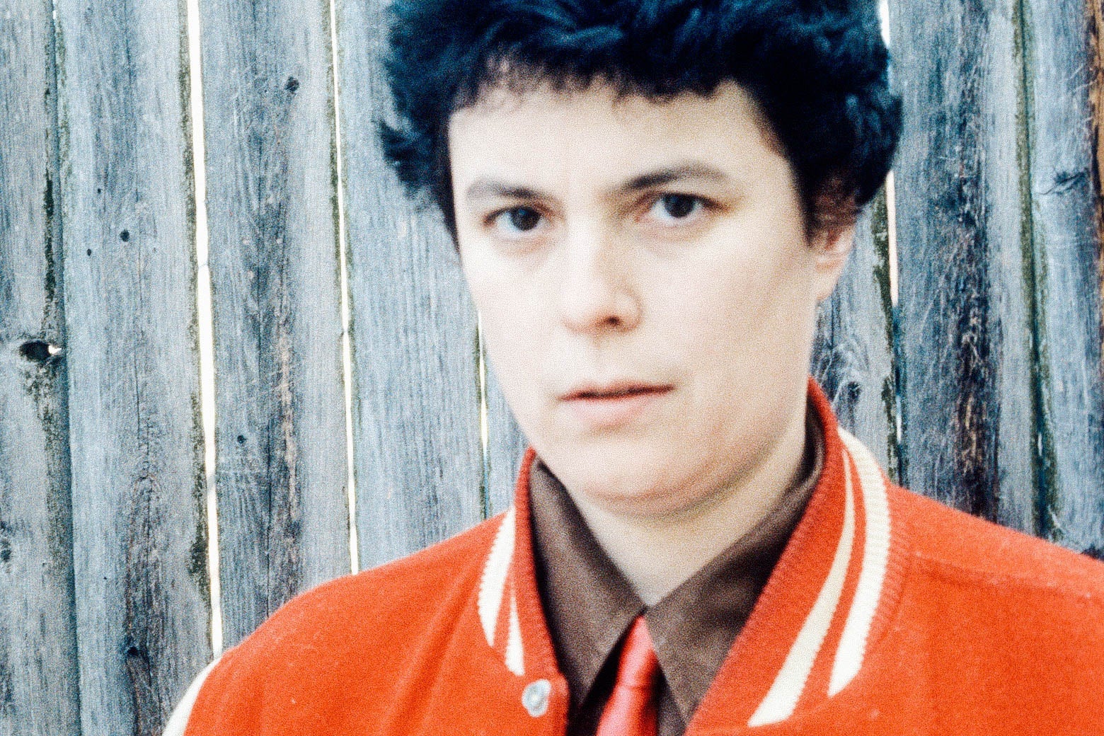 A young woman with short hair, a letter jacket, and a red tie, stares confidently into the camera.