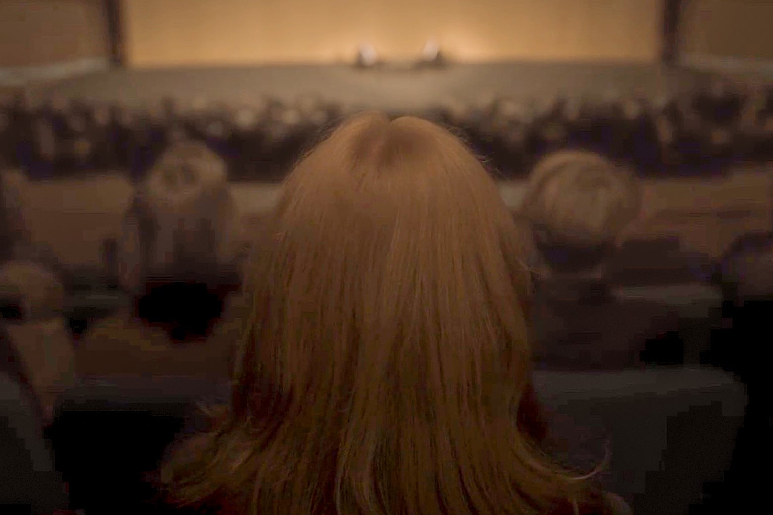 A red-headed woman, facing away from the camera, sits in the audience of a theater.