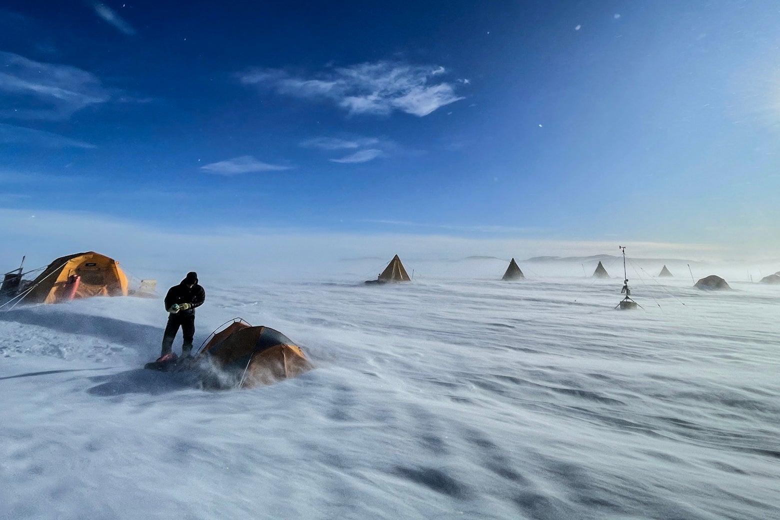 A person stands next to a tent in a landscape covered in snow, five more tents can be seen in the background.