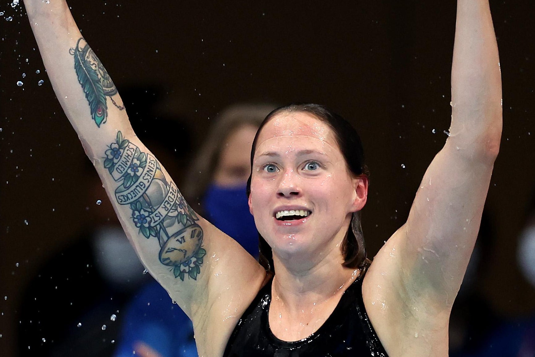 Kohler smiling with her arms raised in her swimsuit, a large tattoo described below visible on her right arm