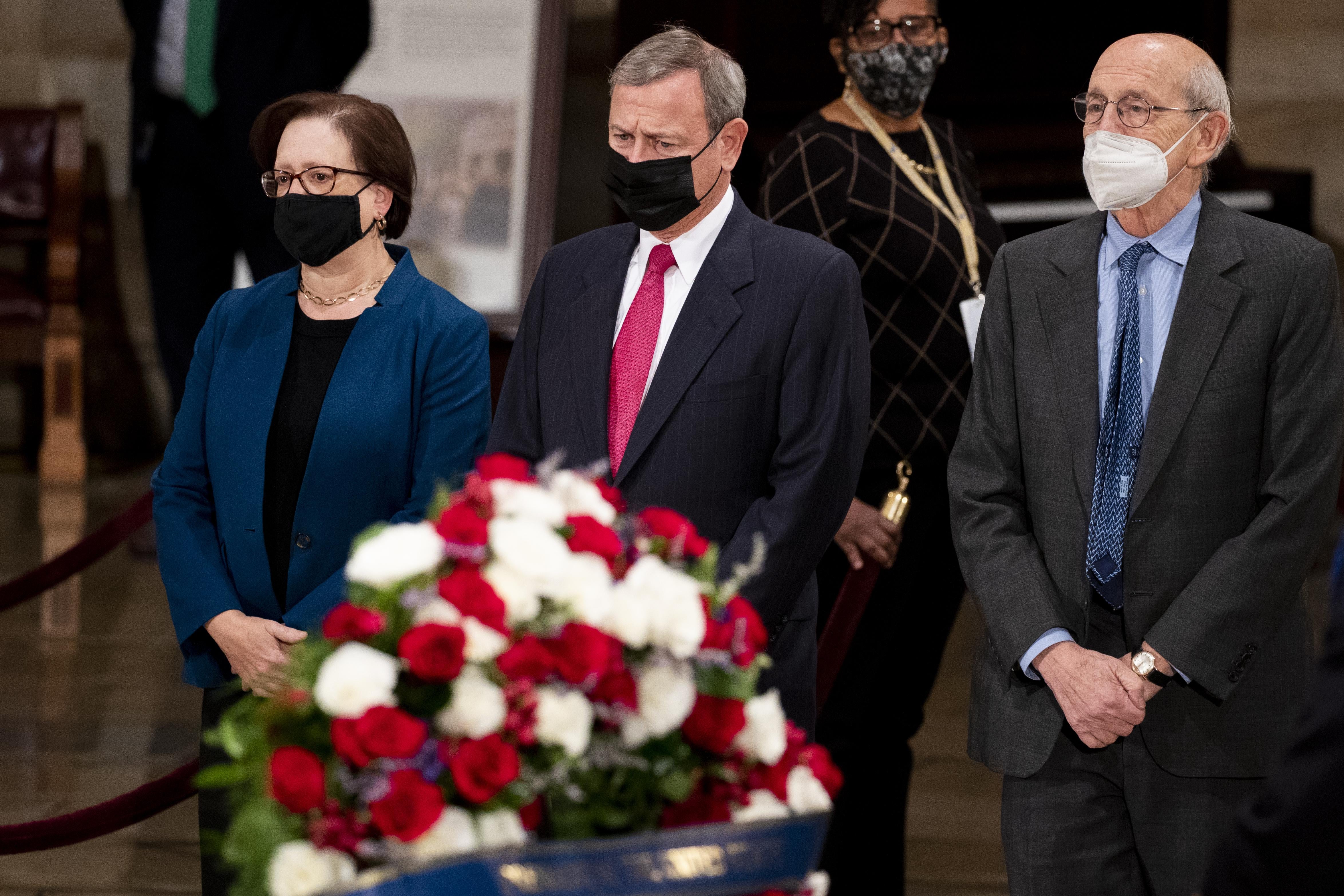 The three justices standing side by side with their arms clasped in front of themselves, standing in front of flowers and the casket of Bob Dole, which is out of the shot.