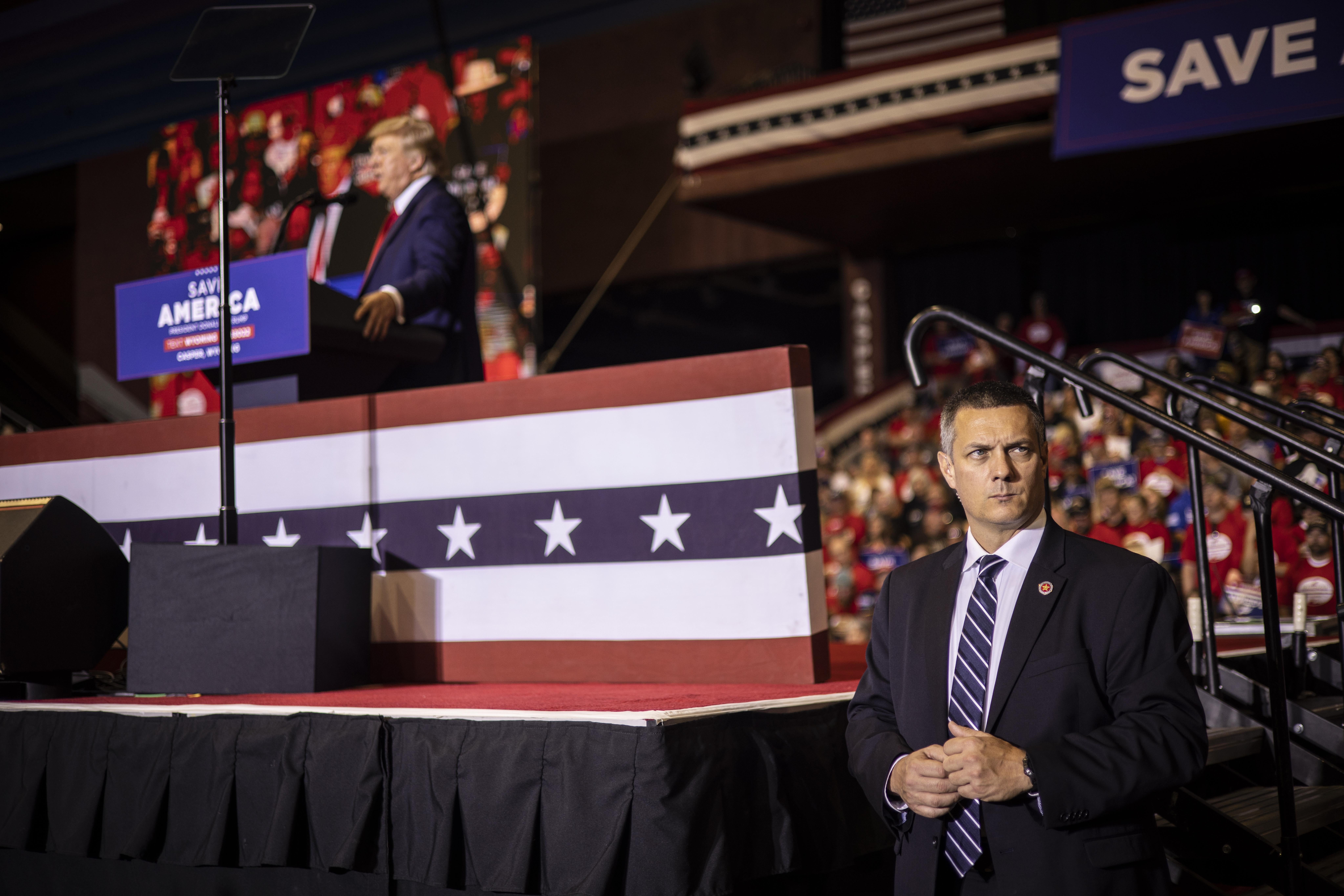 A serious-looking man in a suit stands guard below the stage where Trump speaks at a lectern in front of a crowd
