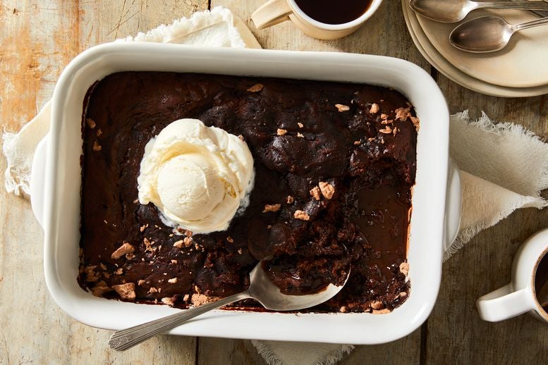 Molten chocolate cake in a ceramic baking dish next to mugs and plates on a wooden table. There's a spoon scooping into the cake and a scoop of vanilla ice cream on top.