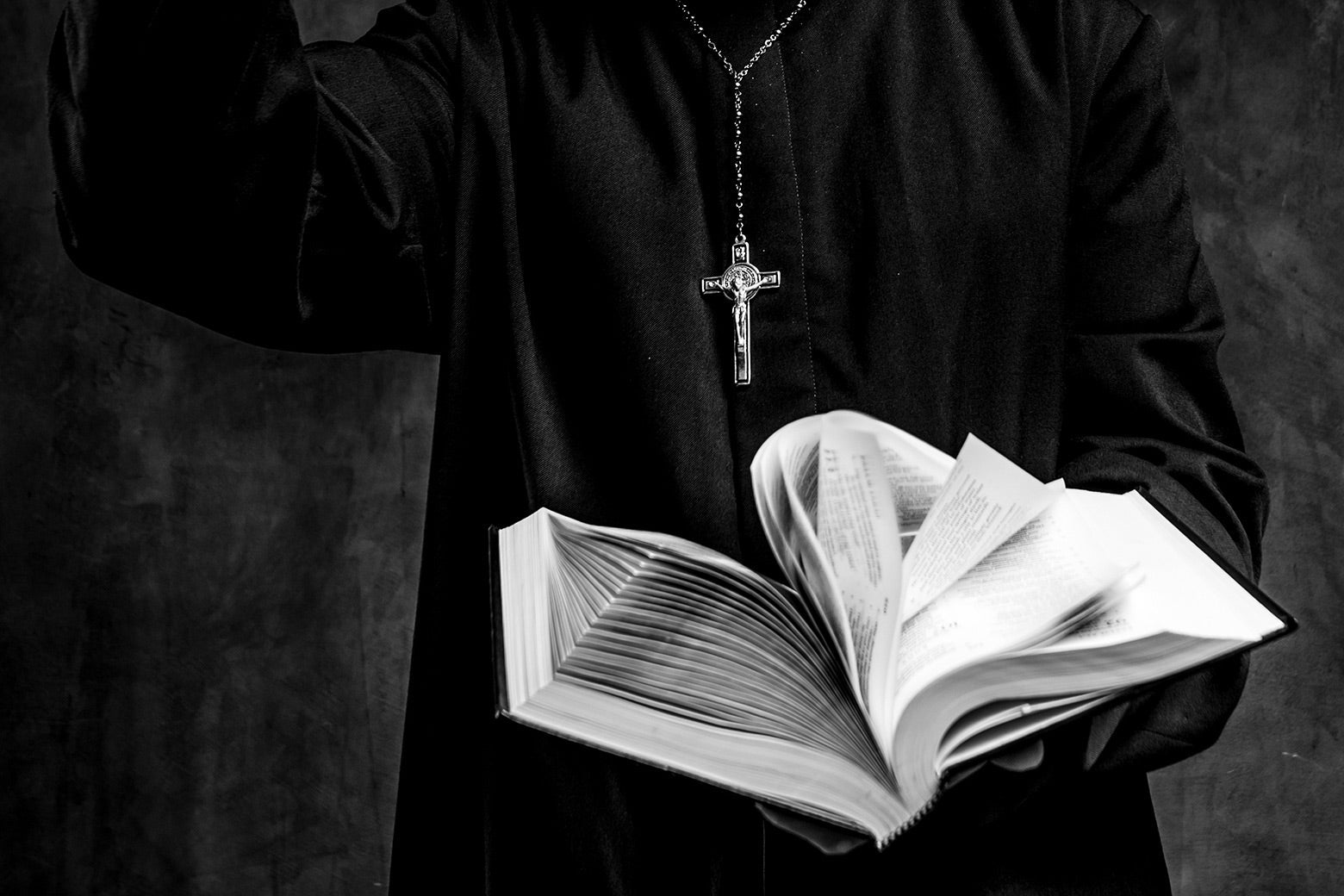 A priest in a black cassock with a crucifix around his neck raising his right arm and holding a Bible open in his left hand