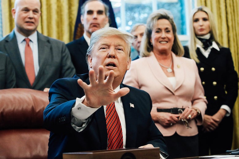Donald Trump, seated at his desk, reaches out a hand toward the camera. People stand behind him.