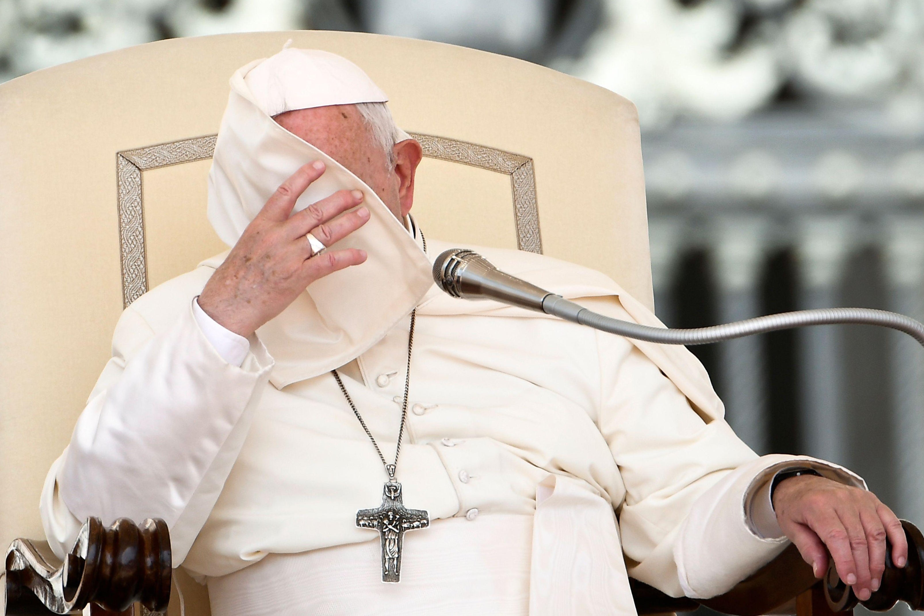 Pope Francis' reaches his hand up as the corner of his cape pellegrina covers his face while he sits in a chair.