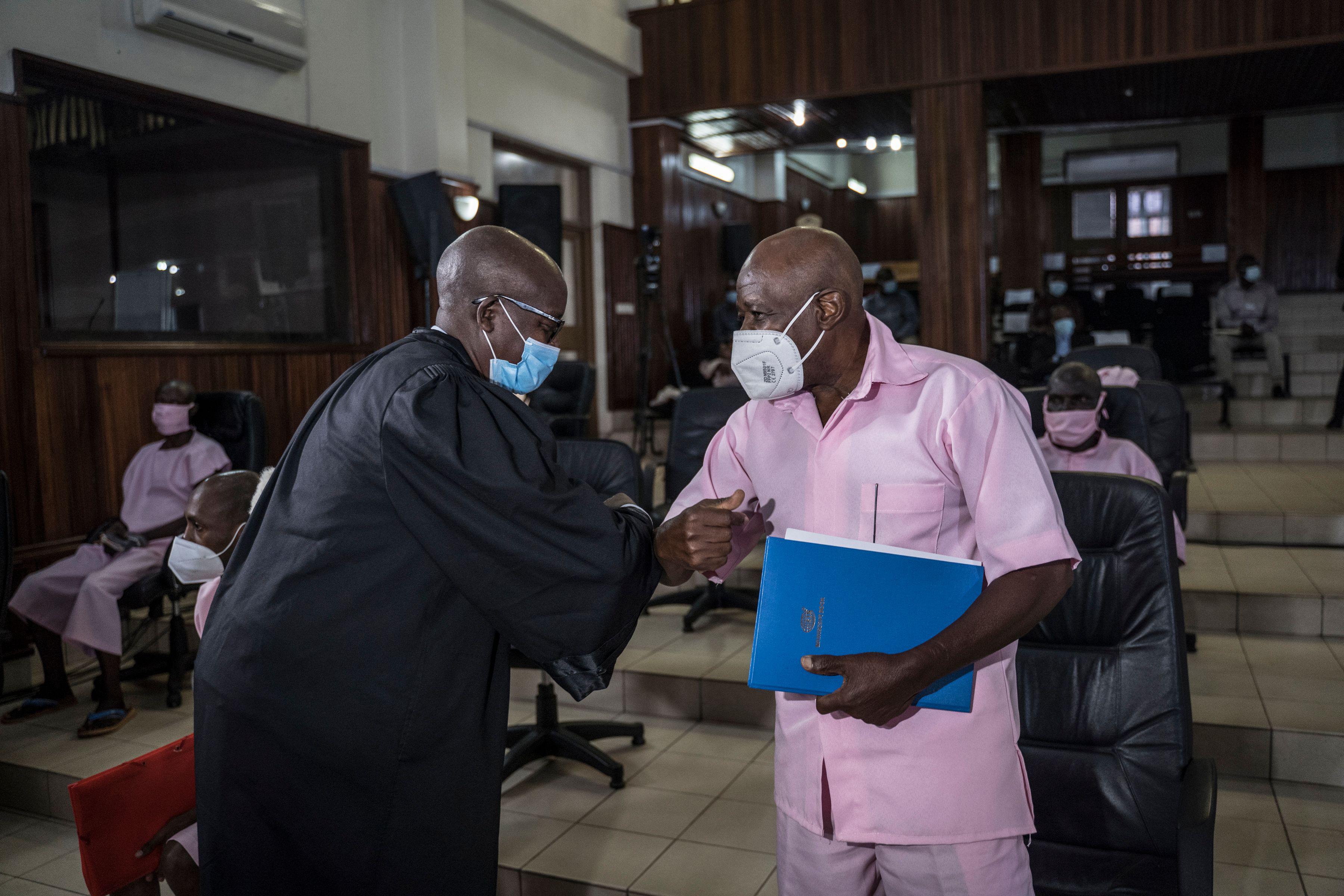 A man in black robes and a mask elbow-bumps with a man in pink clothes and a mask in a courtroom.
