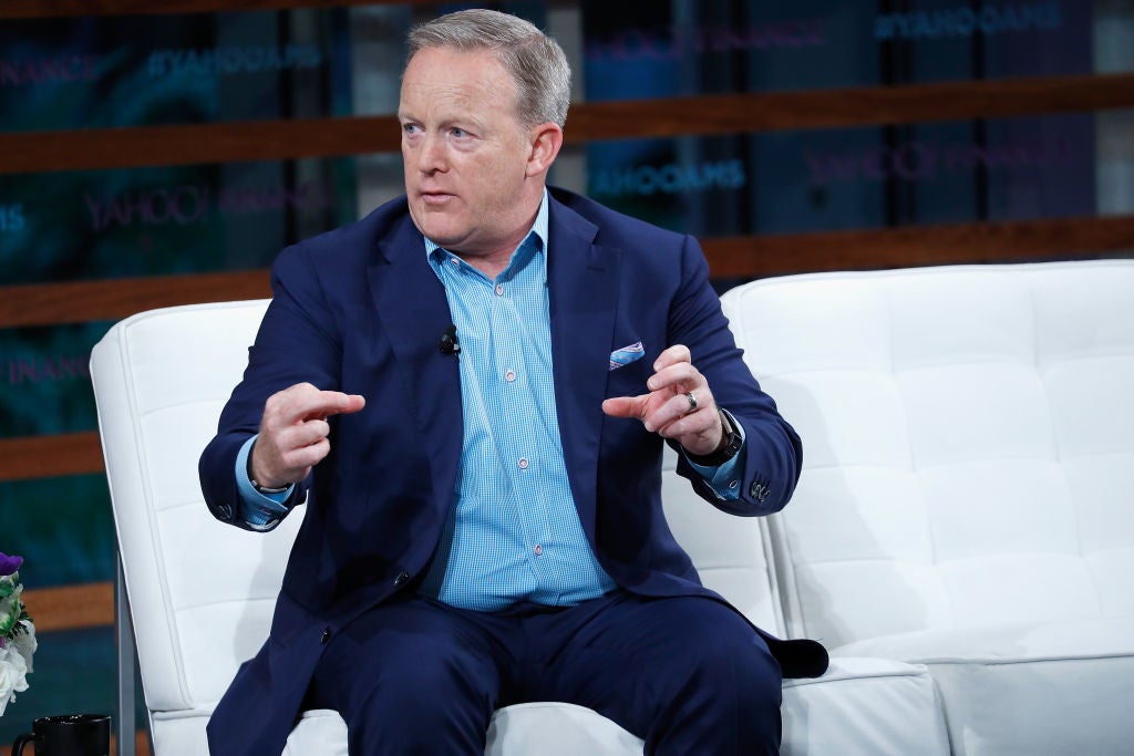 Sean Spicer makes a grasping motion with both hands while seated on a white couch on a stage.