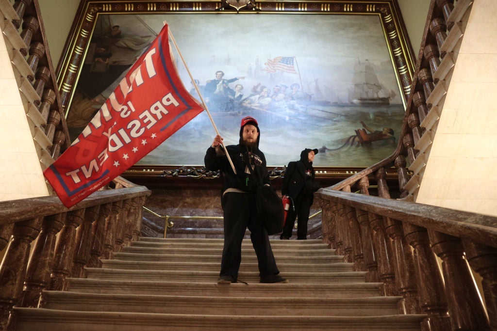 A man carries a large Trump flag up a staircase in front of a large painting.