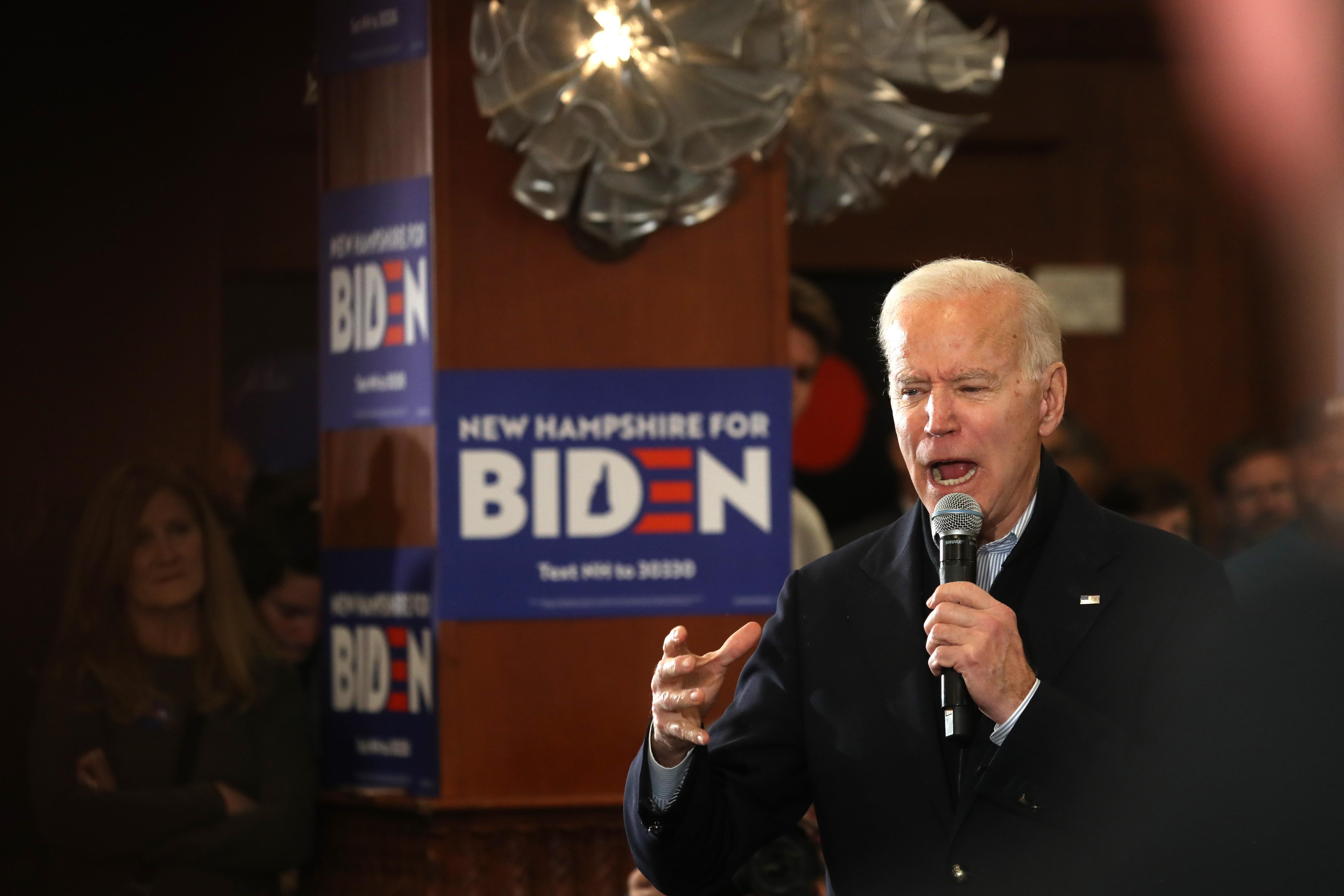 Joe Biden holding a microphone, mid-word, in front of a Biden campaign sign.