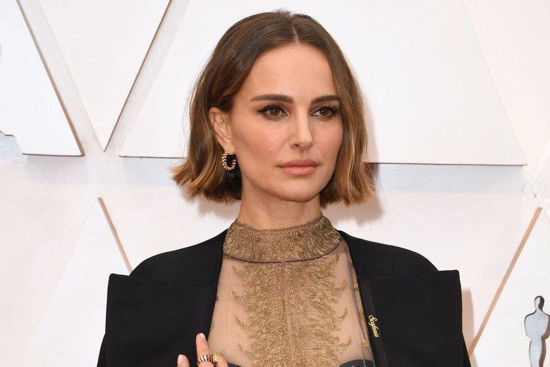 Natalie Portman on the red carpet. She is wearing a black cape with names written on it in gold.