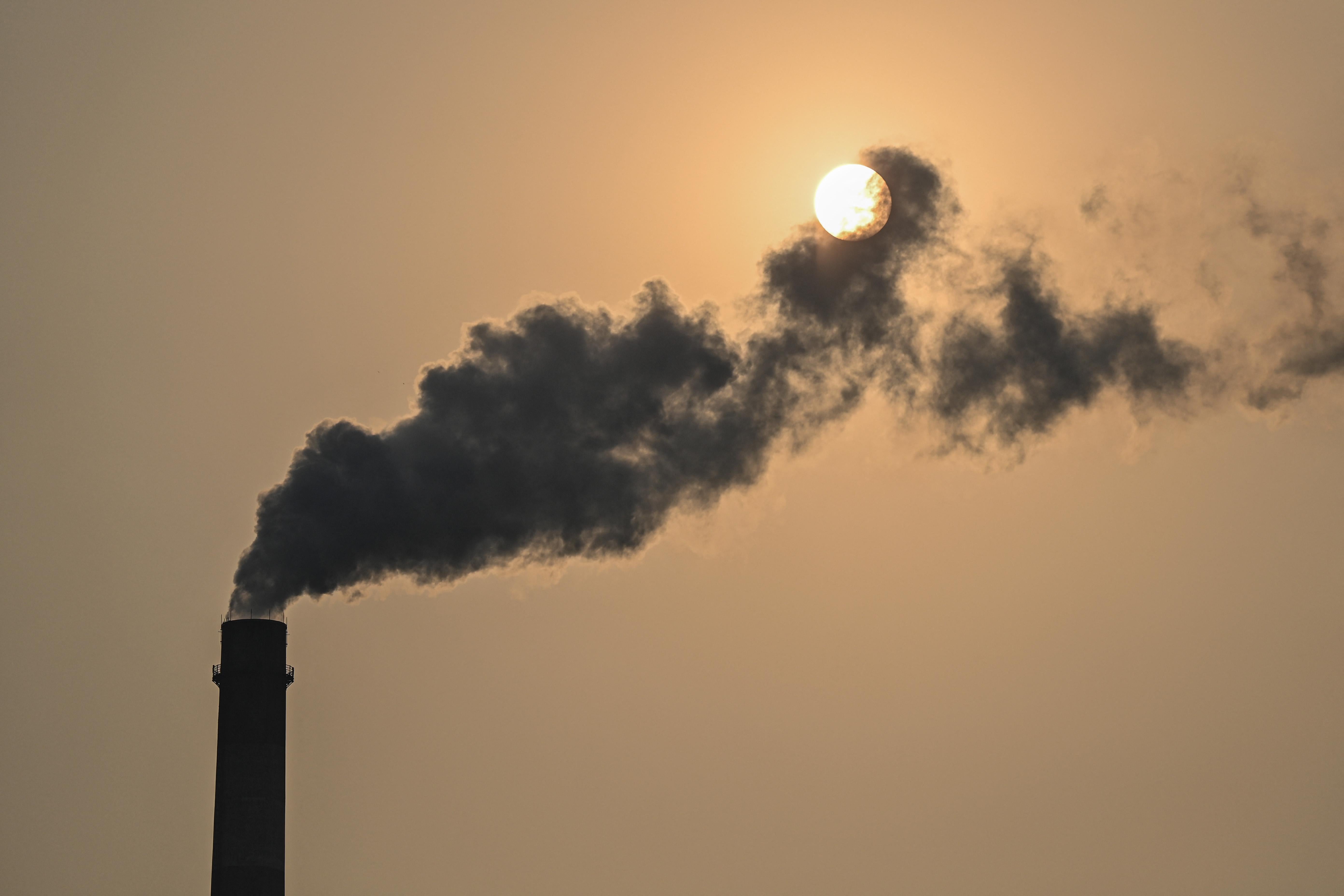 To the left of the image, a smokestack  releases a tower of smoke into a greyish, polluted sky. The sun can be seen behind the pollution. 