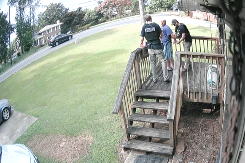 A man being arrested at a suburban home in crocs and shorts and a tee.