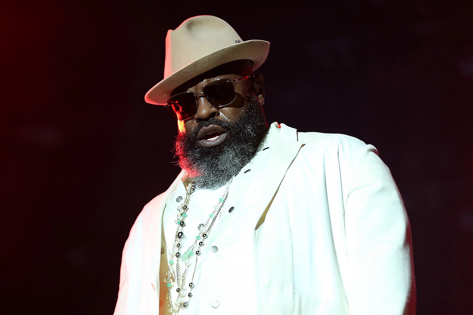 The rapper wears sunglasses, a thick beard, a tan hat cocked to one side, a white suit, and several colored stone necklaces
