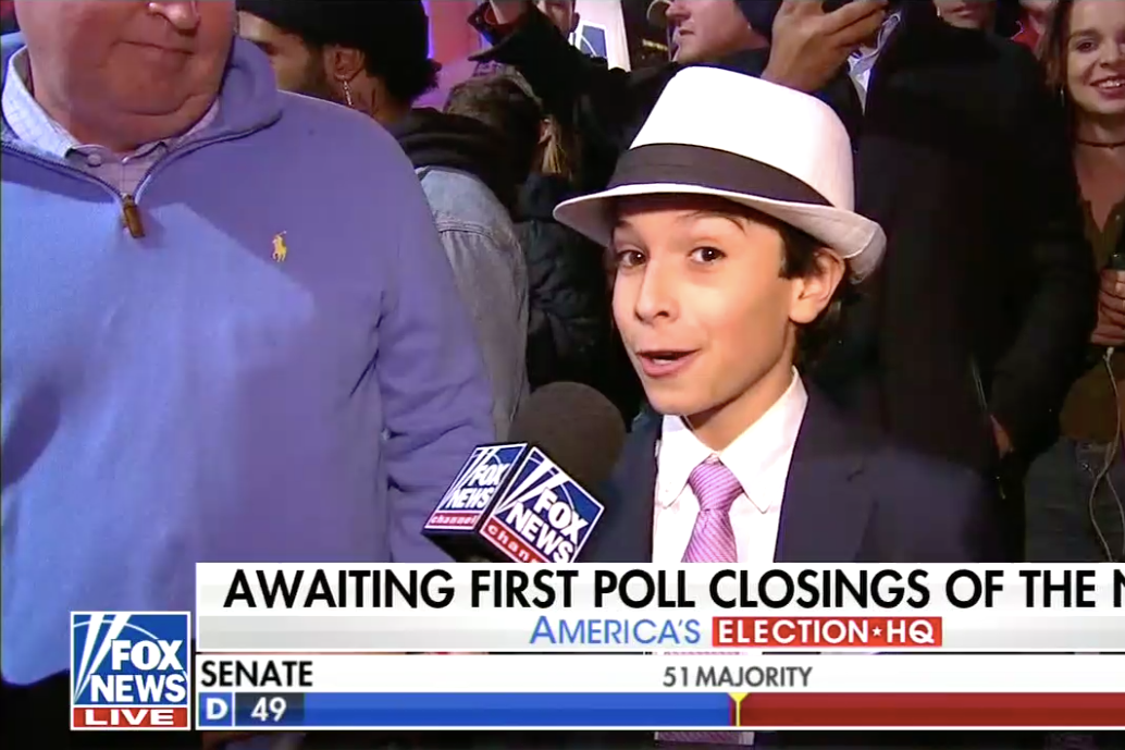 A boy wearing a fedora talks to a Fox News reporter on the street.