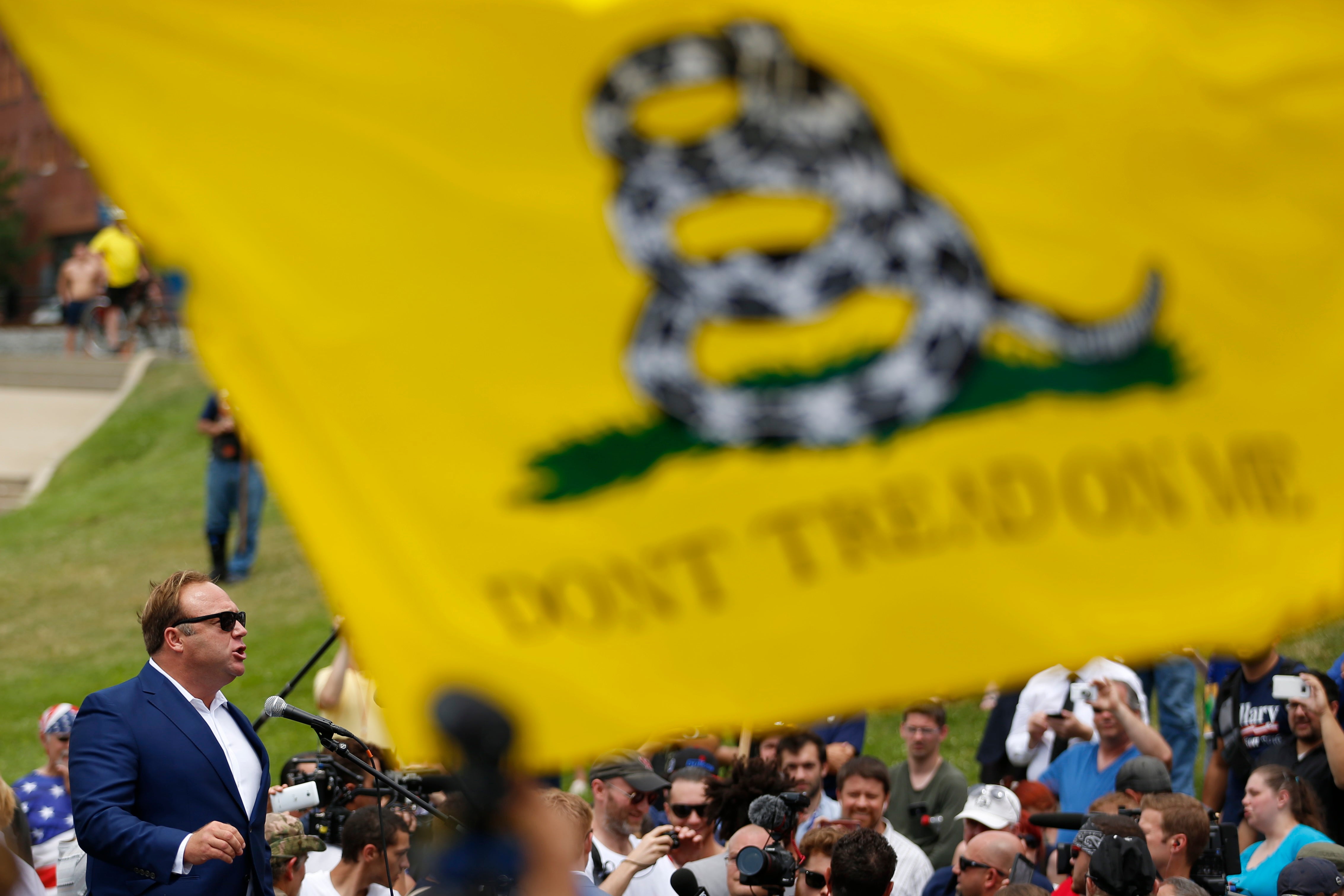 Alex Jones speaks at a public event behind a "Don't Tread on Me" flag.