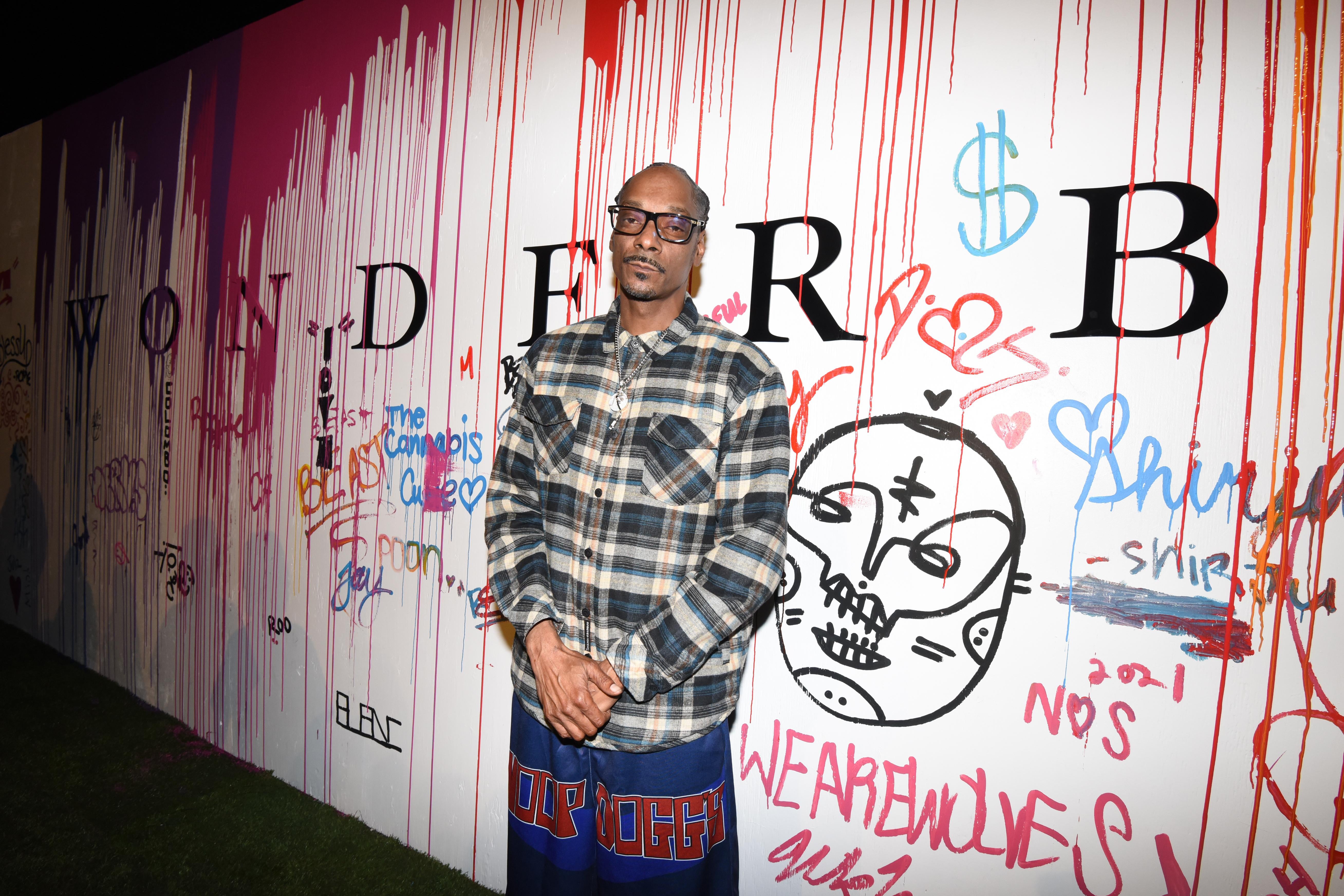 Snoop Dogg stands in front of a heavily graffiti'd white wall.