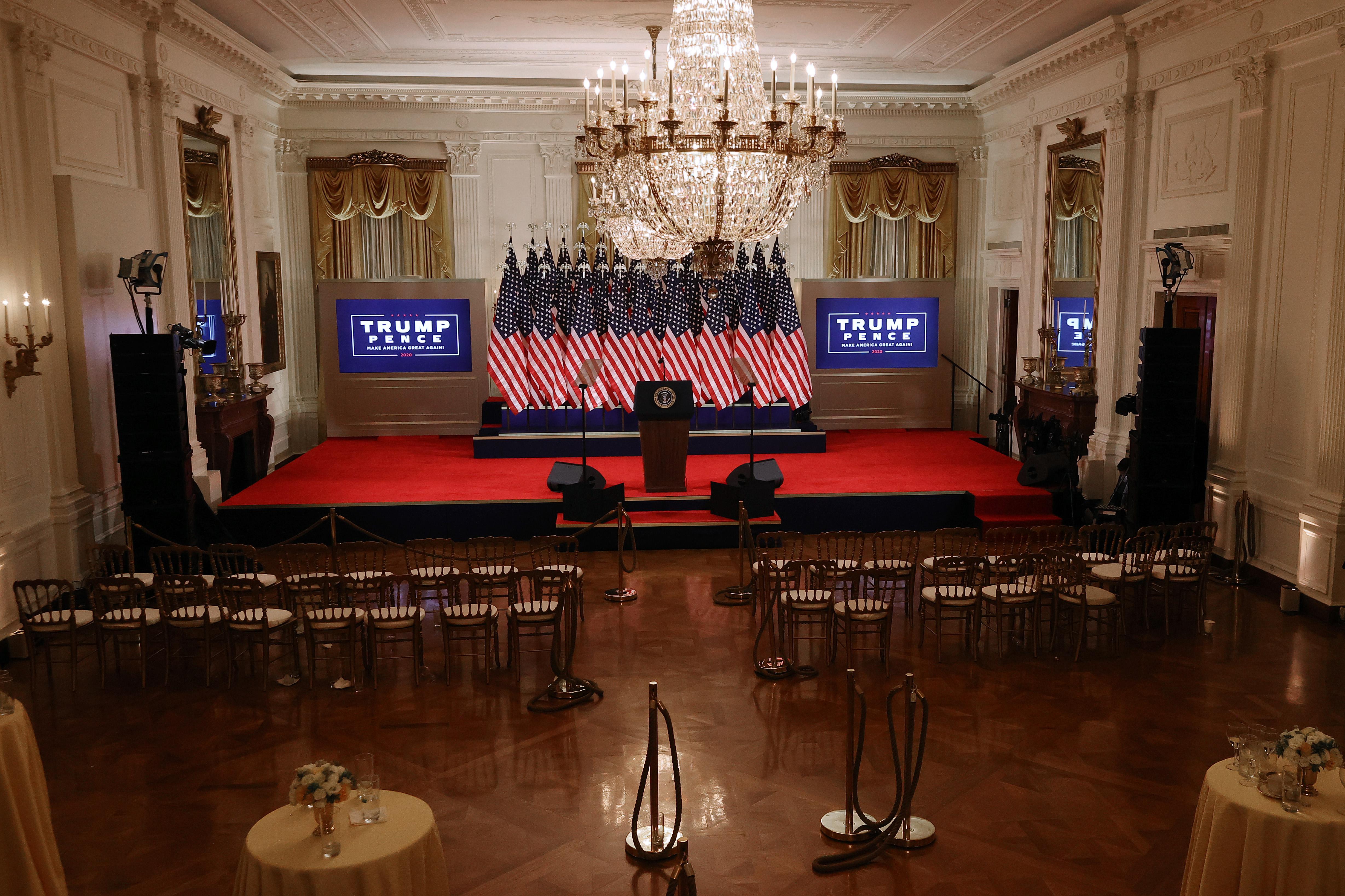 A room empty of people, with rows  of vacant chairs facing a presidential lectern on a red-carped stage, backed by a solid mass of American flags on flagpoles and flanked by Trump-Pence logos on screens. 