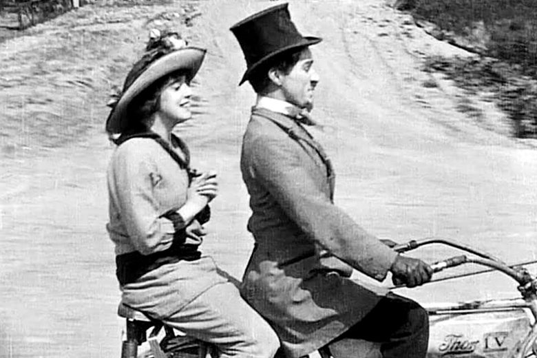 Mabel Normand on the back of a motorbike that Charlie Chaplin drives