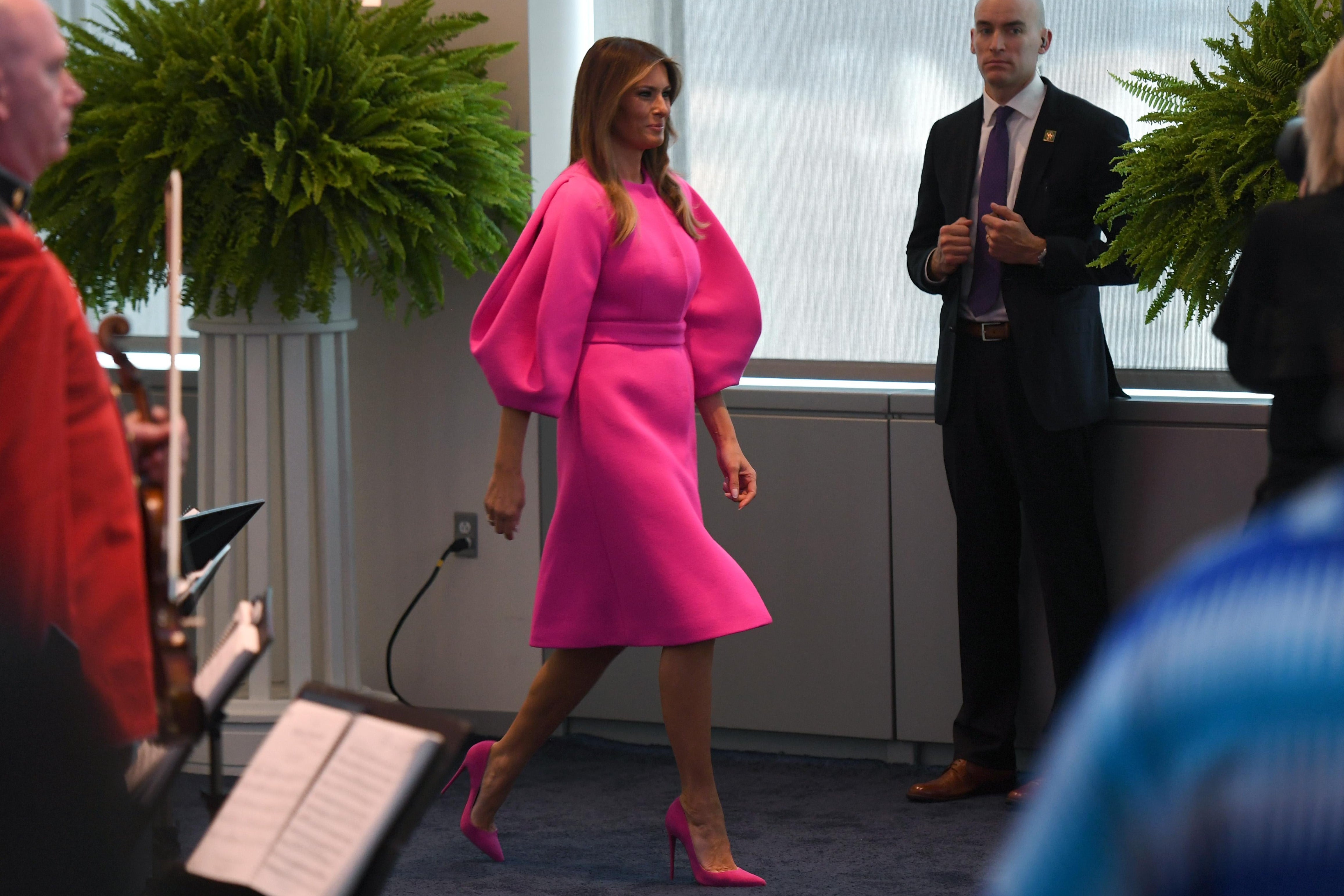 Melania Trump walks past a fern, as several people look on, wearing a hot pink dress with puffy long sleeves and matching heels.