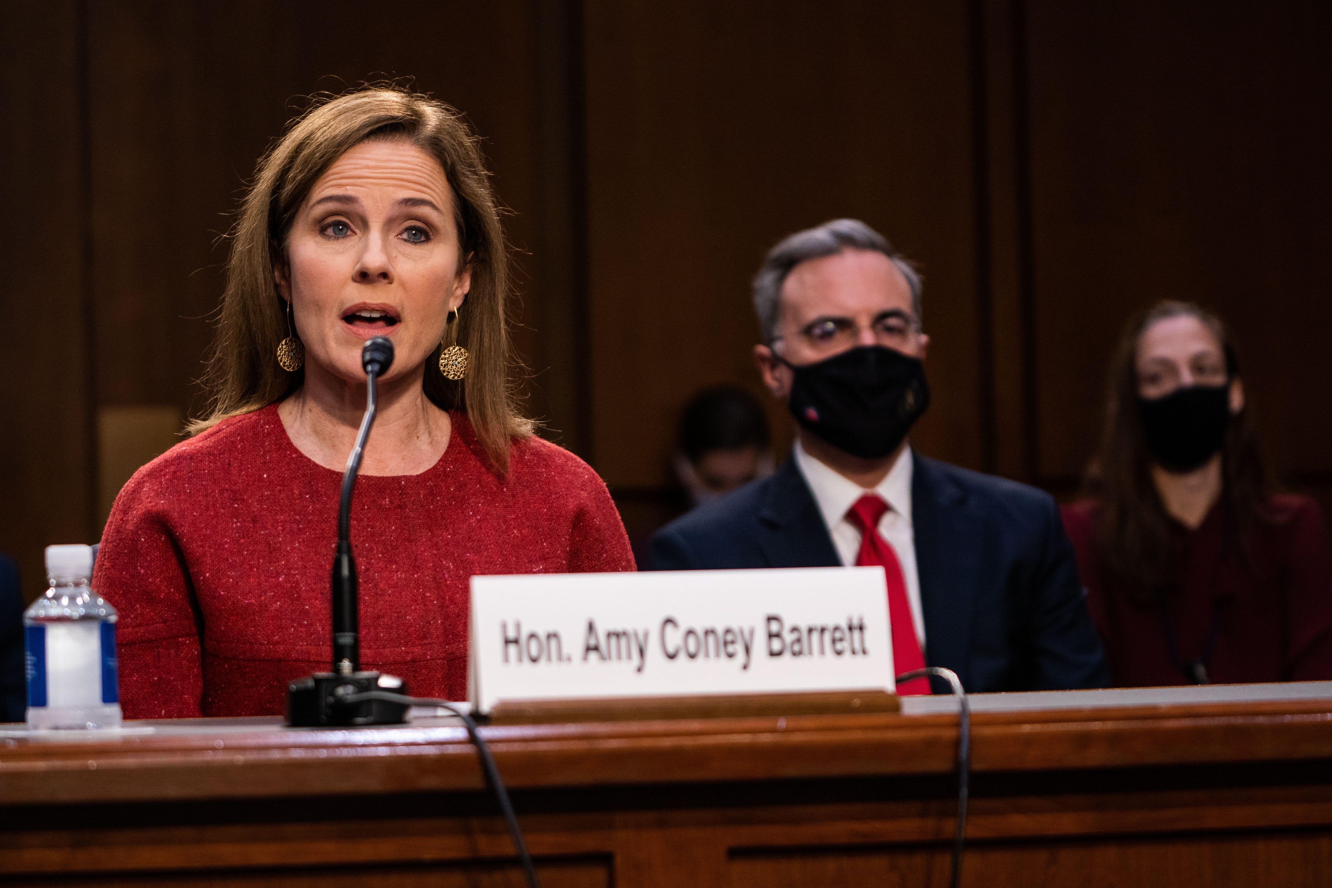 A woman in a red sweater speaking into a microphone sits behind a nameplate that says, "Hon. Amy Coney Barrett."