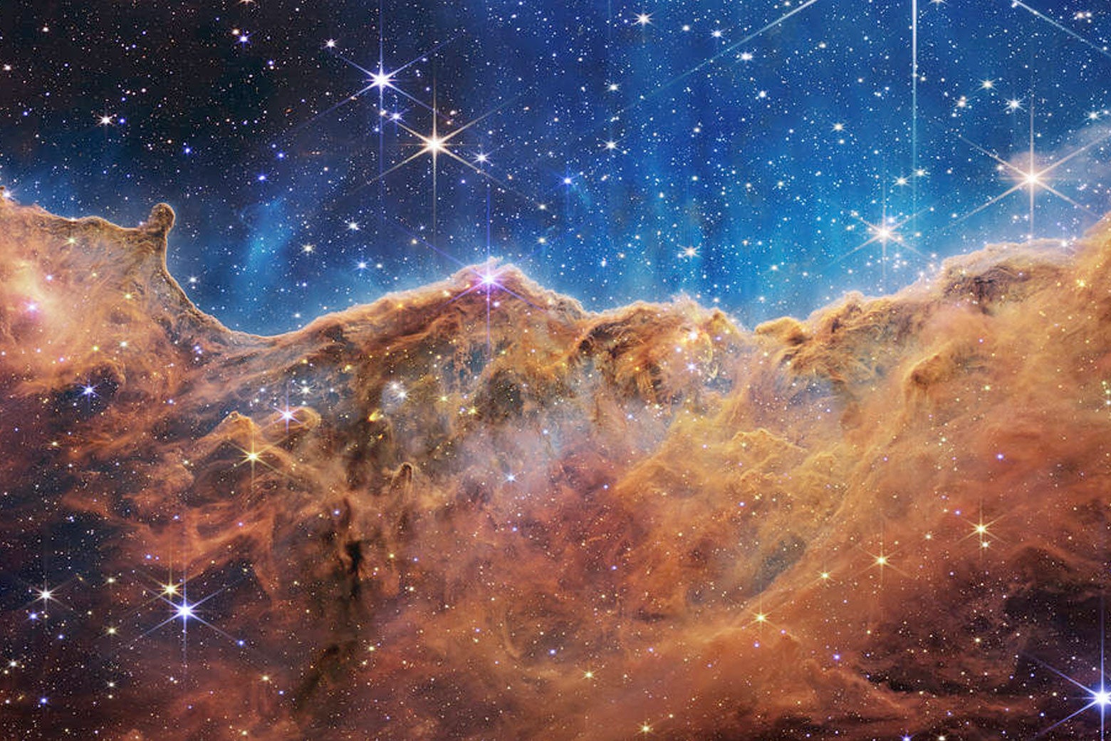 A blue sky in outer space speckled with stars. Giant plumes of orange-red gas cover half the frame. They look like fluffy, bejeweled mountains. The image evokes the word "majestic."