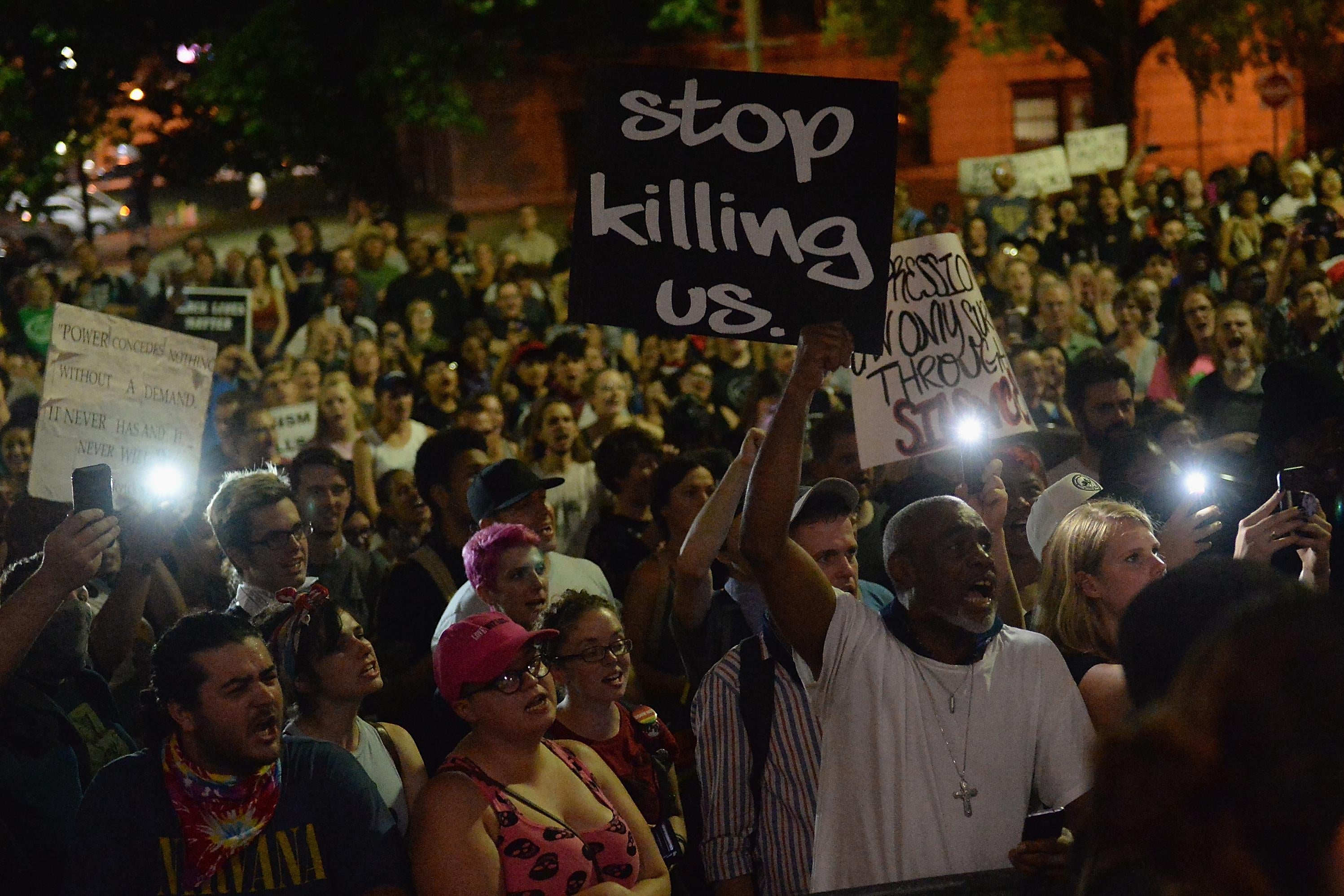 A few people hold lights from their cell phones at a night-time protest. A man in a white tee shirt at the front holds a sign that says, "stop killing us."
