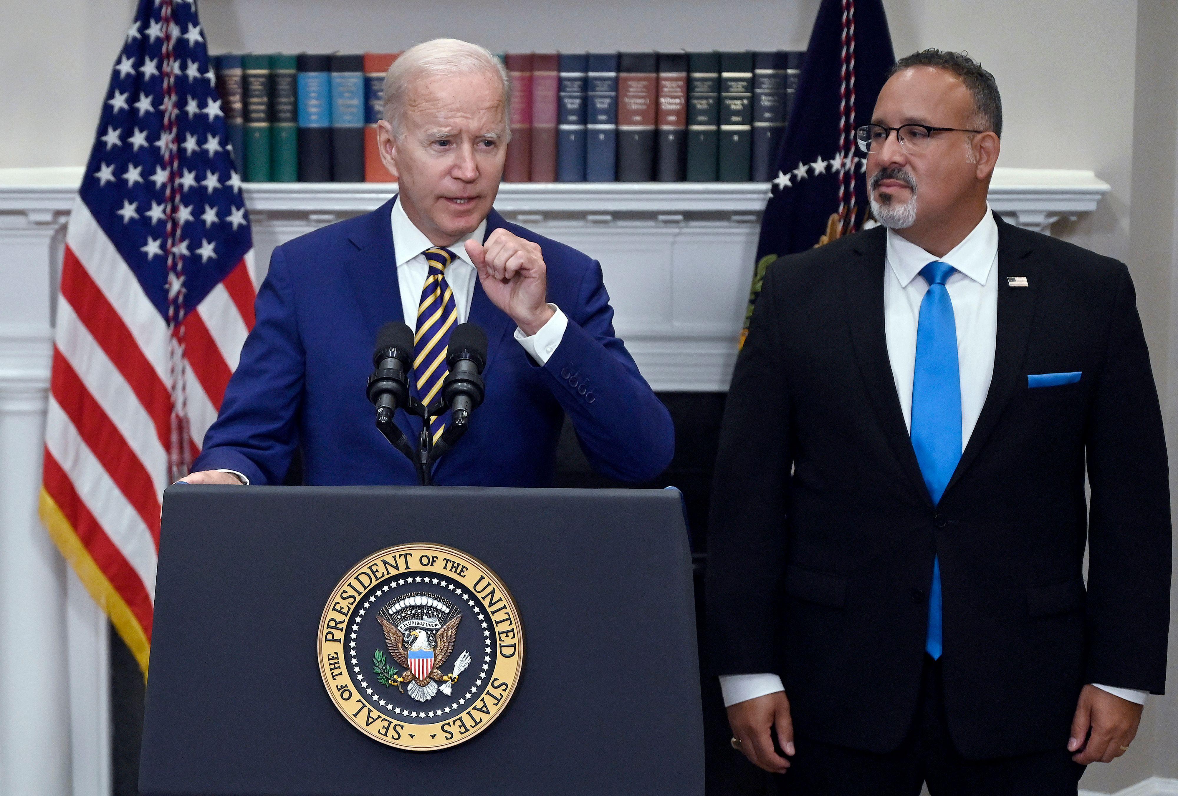 Biden speaks at a podium while Cardona looks on.