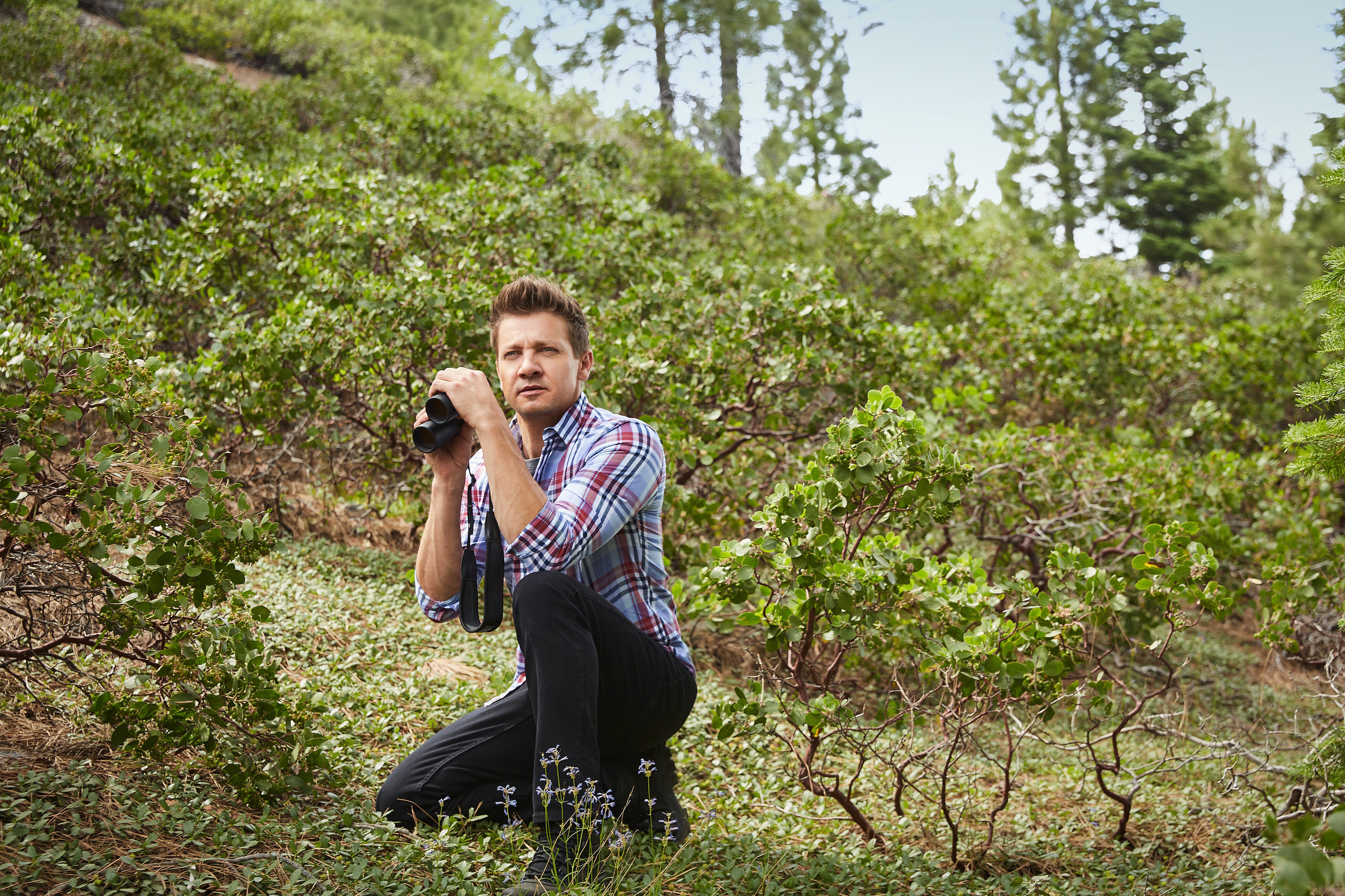 Jeremy Renner crouches in the wilderness, holding a pair of binoculars but not looking through them.