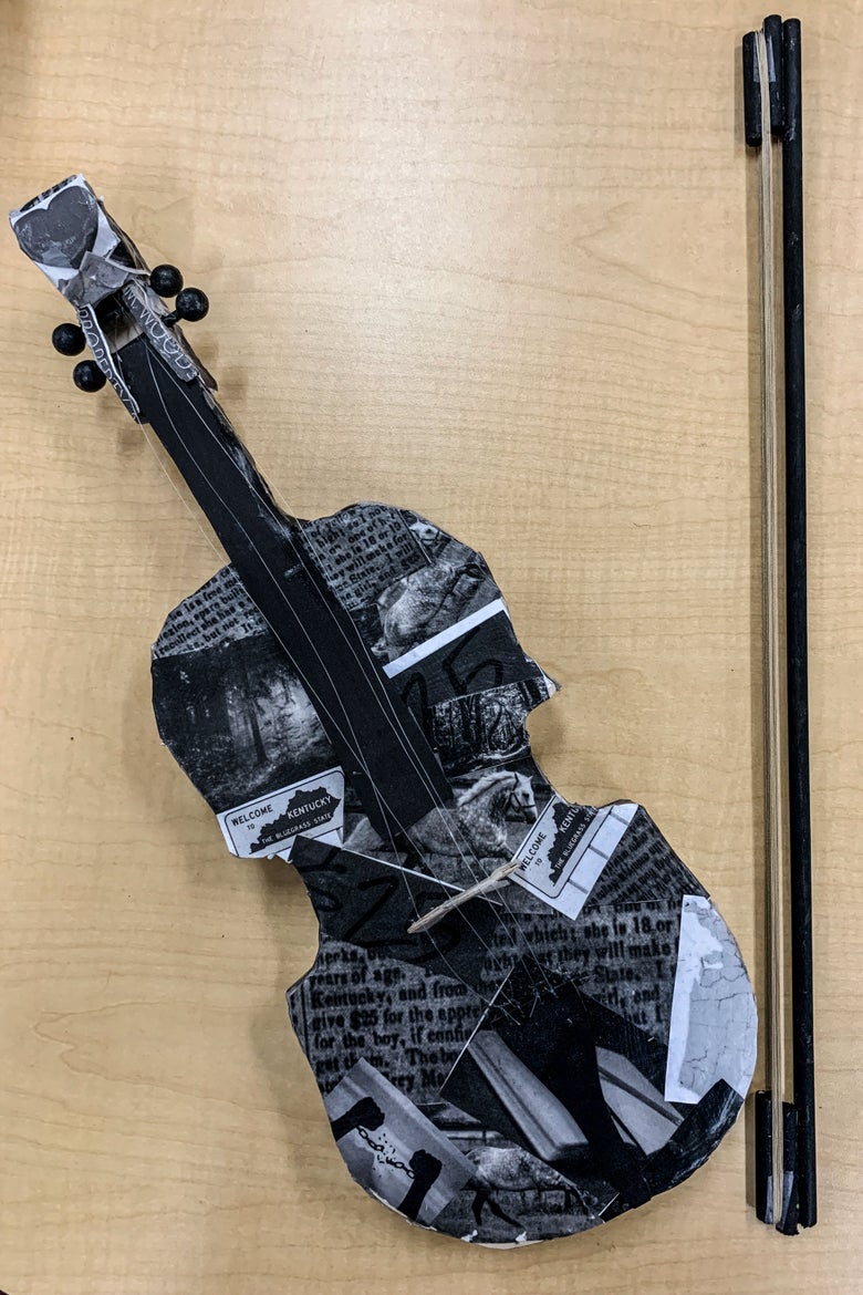 A black cardboard fiddle covered in newspaper clippings and other cutout images with a bow sitting beside it on a table