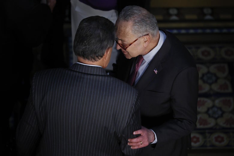 Two men in suits speak, their heads close together, one touching the other on the upper arm.